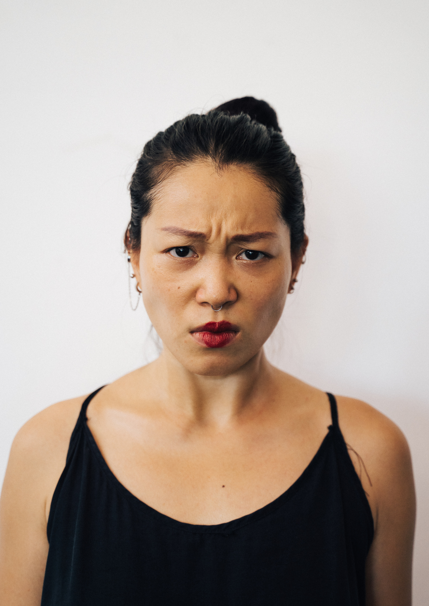 A woman with dark hair in a bun, wearing a black tank top, making a confused or skeptical facial expression with furrowed brows and pouty red lips, against a plain white background.