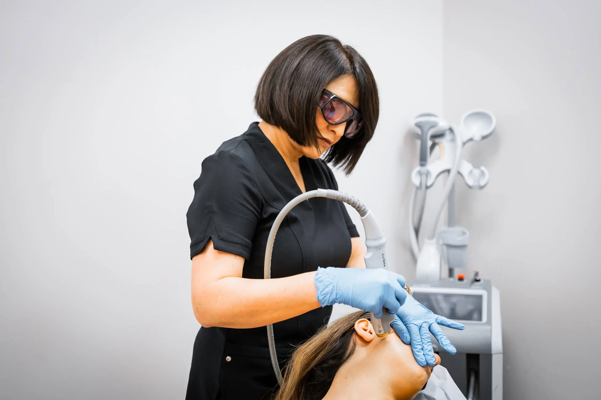 A medical professional in black attire, wearing safety glasses and blue gloves, performing laser treatment on a woman's face in a clinical setting.