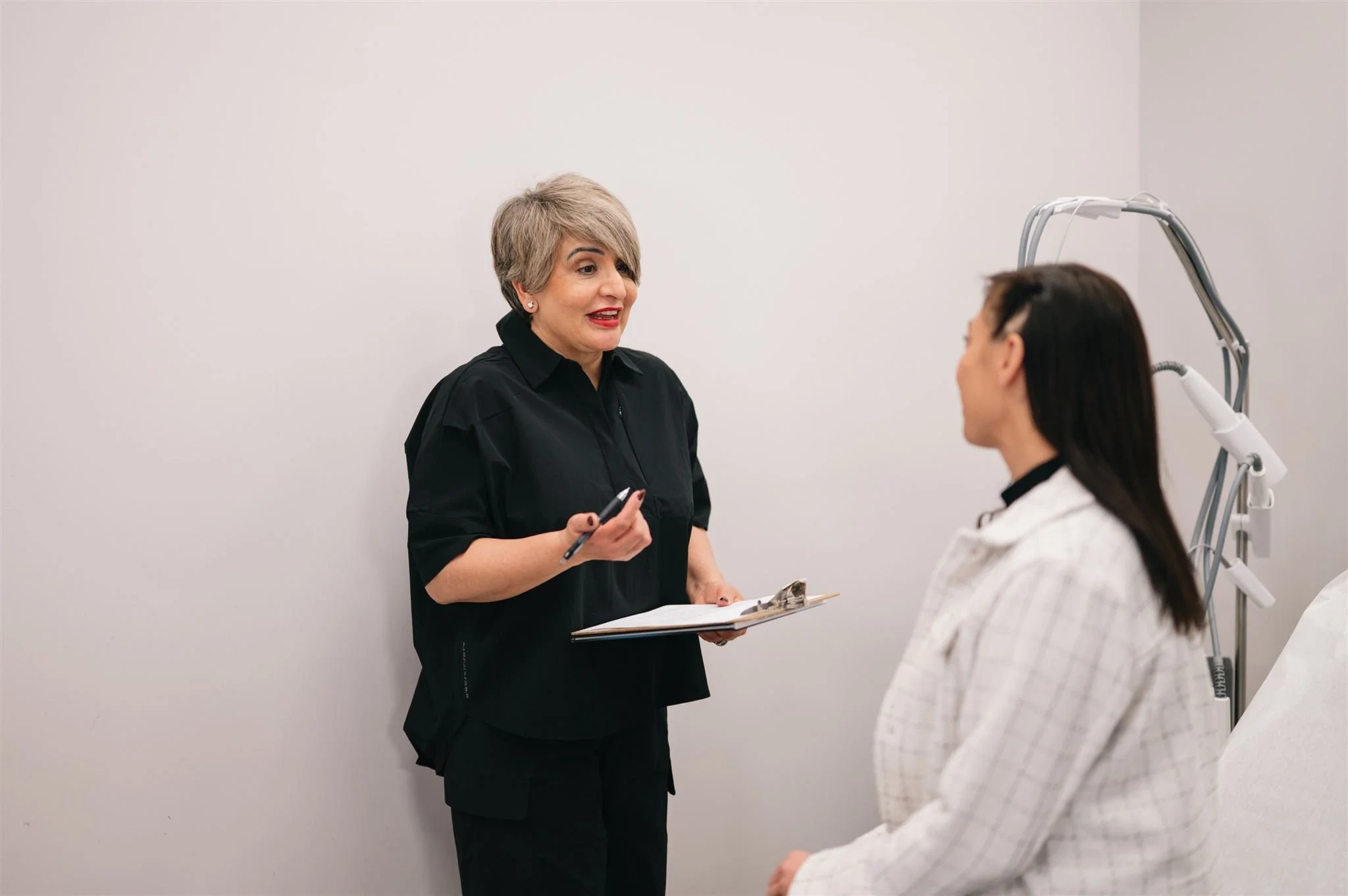 A doctor and a woman in a consultation room, with the doctor holding a clipboard and speaking to the woman, who is sitting on a medical bed.