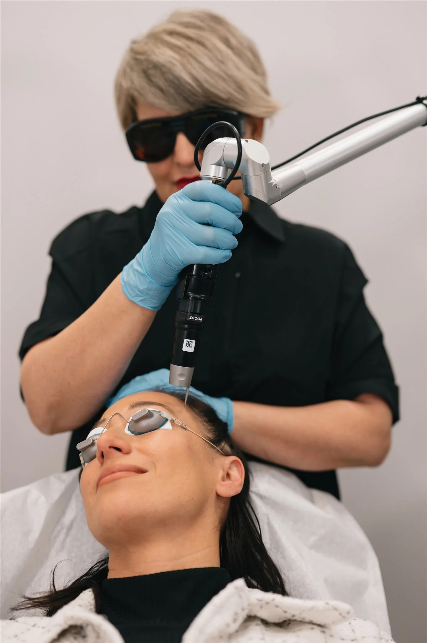 A woman lying down with protective eyewear receives a cosmetic cosmetic treatment from a professional wearing gloves and sunglasses, using a laser device.