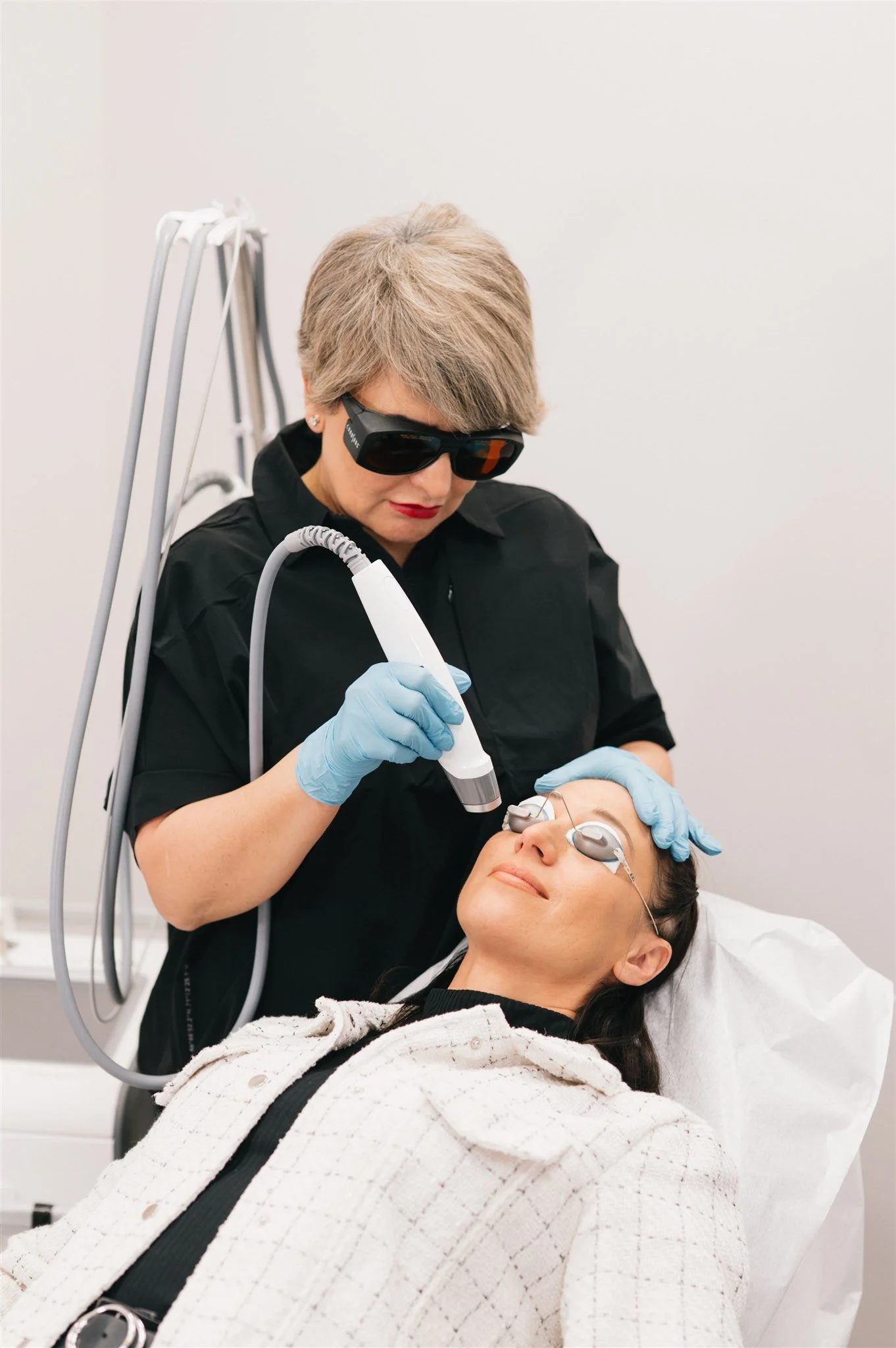 A woman undergoing a laser skin treatment at a skincare clinic, lying on a treatment bed with protective eye goggles, while a technician in black attire and gloves performs the procedure using a handheld laser device.