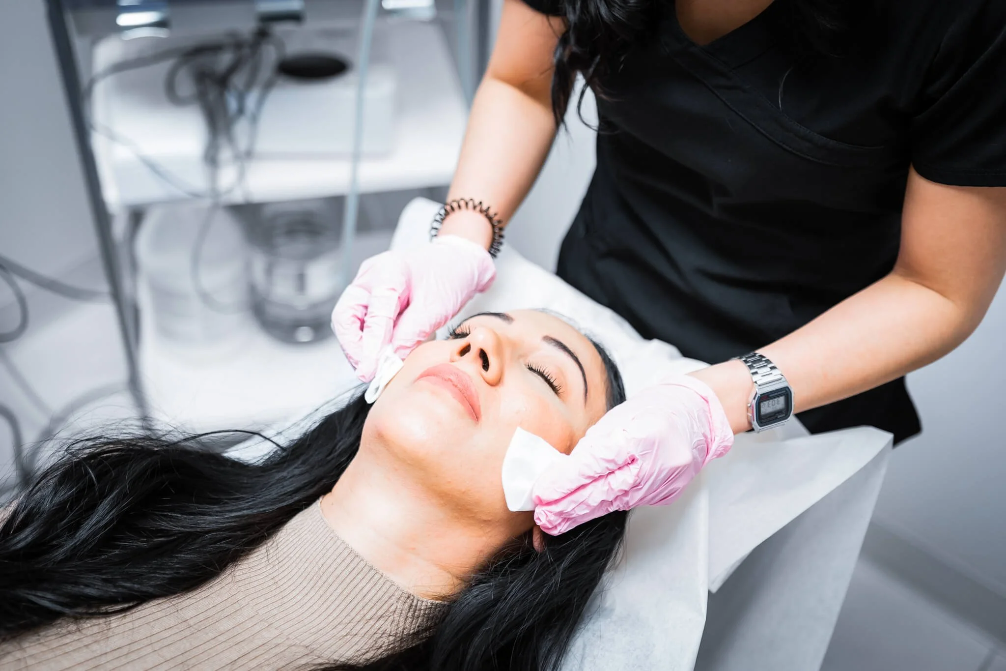 A woman receiving a facial treatment at a spa or clinic, lying on a treatment bed with eyes closed while a professional wearing pink gloves applies a facial mask or cleanser.