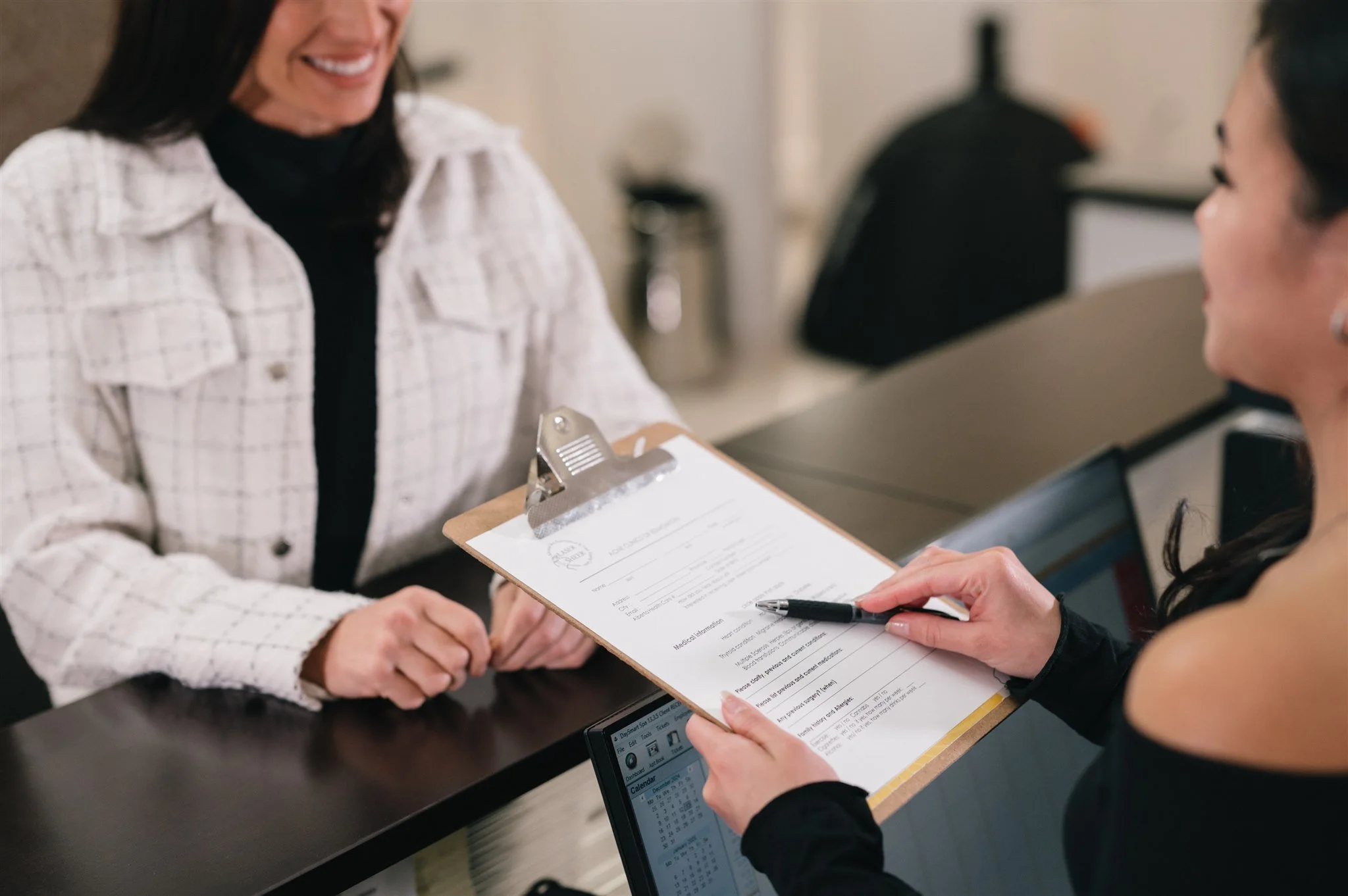 A woman at a reception desk holding a clipboard and pen, smiling and interacting with a woman behind the counter, who is also smiling. The woman behind the counter is holding a pen and appears to be filling out or reviewing a form.