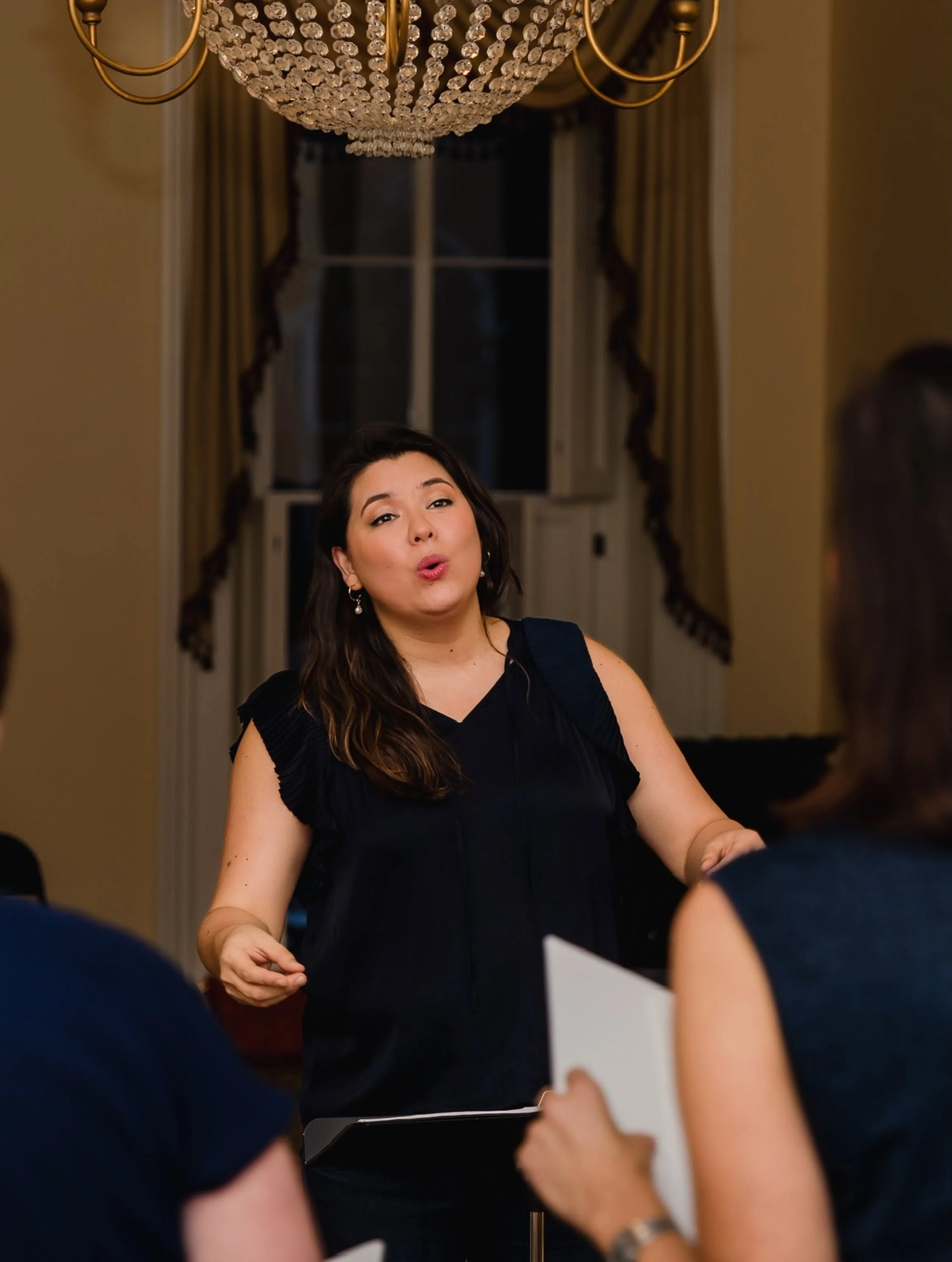 A woman in a black dress is speaking or singing at a formal event, with people seated around her. She has dark hair and earrings, and the background features a grand chandelier, a window with curtains, and warm lighting.