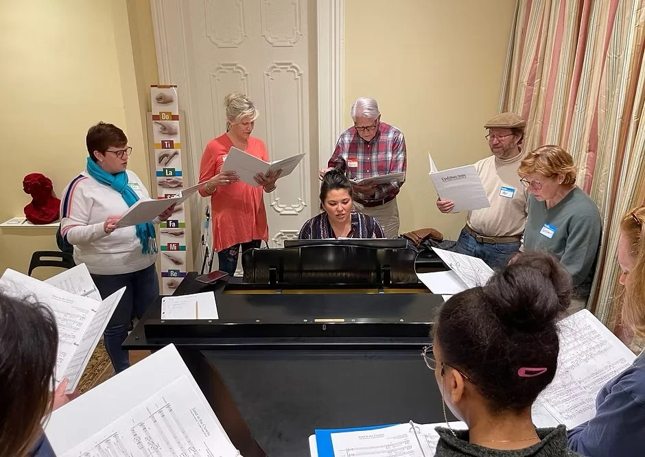 Group of people singing around a piano in a room, some holding sheet music.
