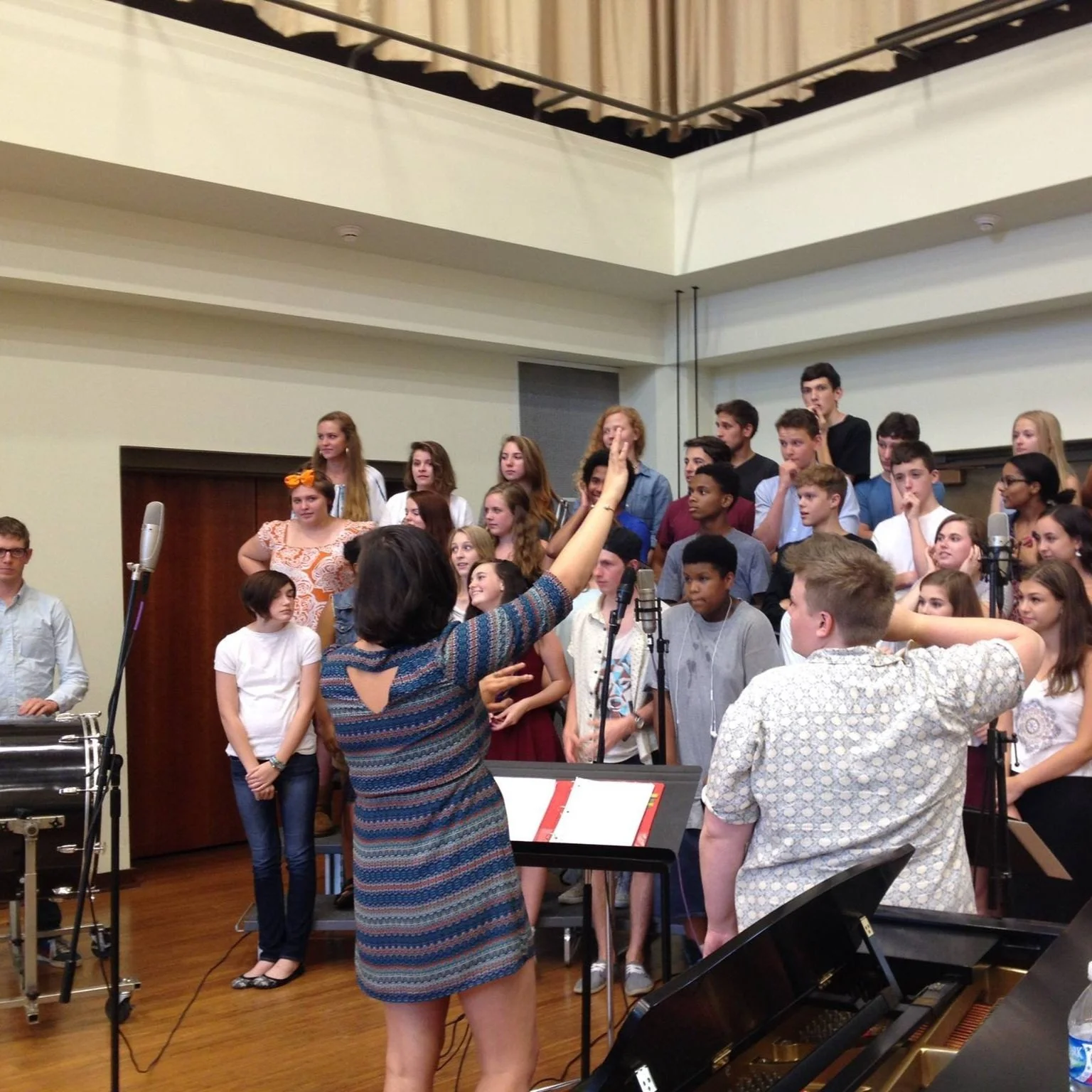A large group of young people on a stage or in a practice room, with a woman conducting or giving instructions, and a keyboard in the foreground.