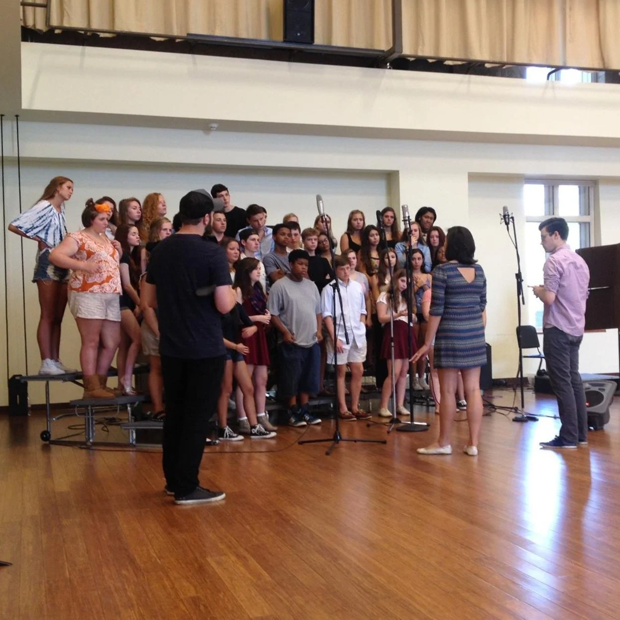A large group of children and teenagers are standing on risers in a music rehearsal room, singing together. There are three adults in front of them, conducting and giving instructions.