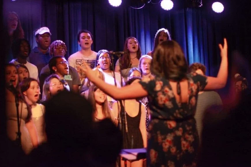 A children’s choir singing with a female conductor leading them, on a stage with a dark curtain and stage lights.