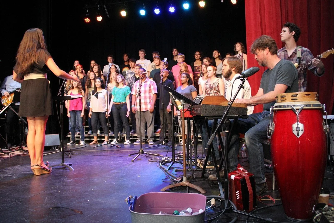 A choir of young people performing on stage with a band. The band has a keyboard player and percussionist, with a conductor leading the choir. Stage lights are above, and red curtains are in the background.