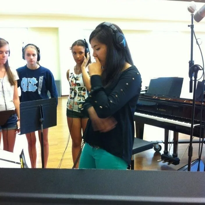 A woman with long black hair in a recording studio, listening to headphones and holding a microphone, with three young girls behind her also wearing headphones, standing near a music stand and a piano.