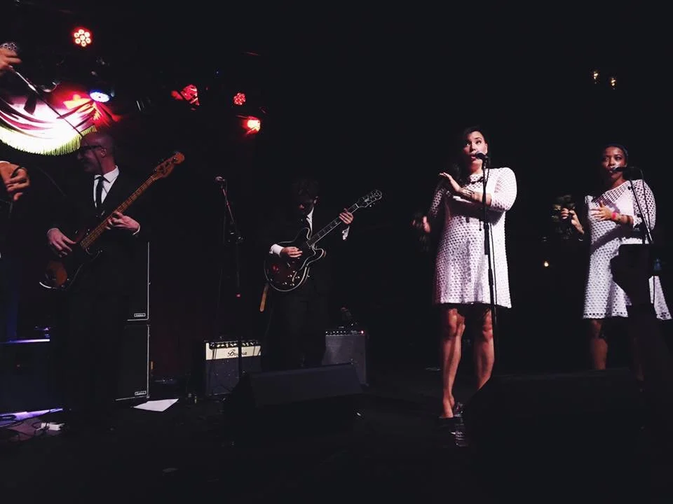 Marianne Cheng singing and playing tambourine with Ernest Ernie and the Sincerities performance at the Bell Hop.

A band performing on stage with three women singing and two men playing guitars, under red stage lights. 