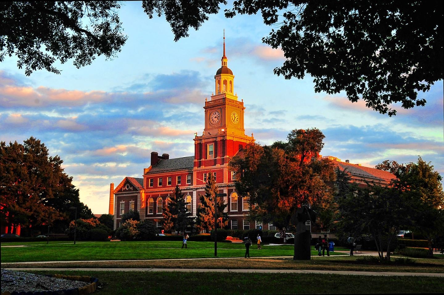 Historical courthouse building with a clock tower, trees surrounding a grassy park area, people walking, and a sunset sky with clouds.