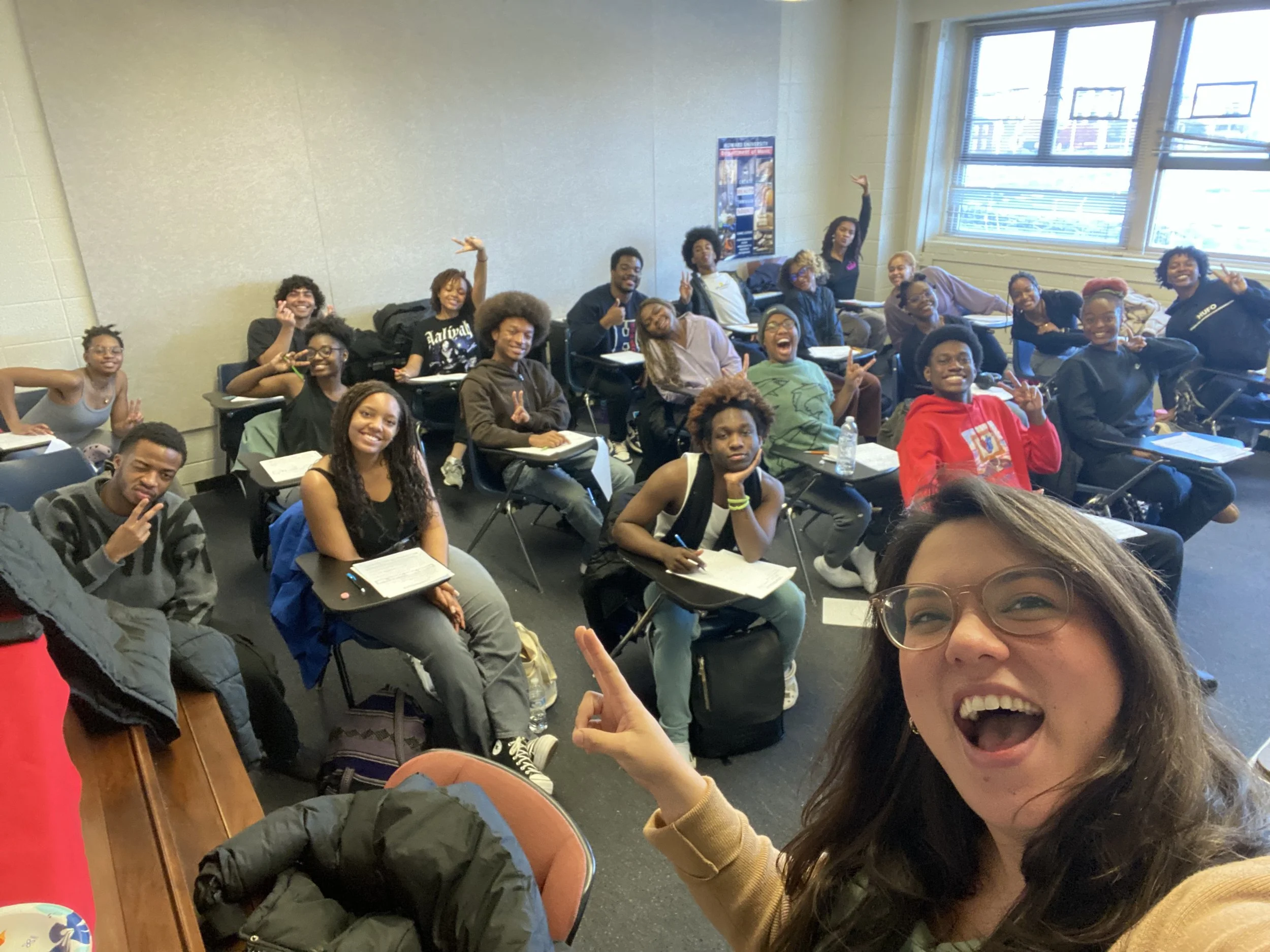 A selfie of a smiling woman with glasses in the foreground and a classroom full of students smiling and making peace signs behind her. The classroom has beige walls and large windows.