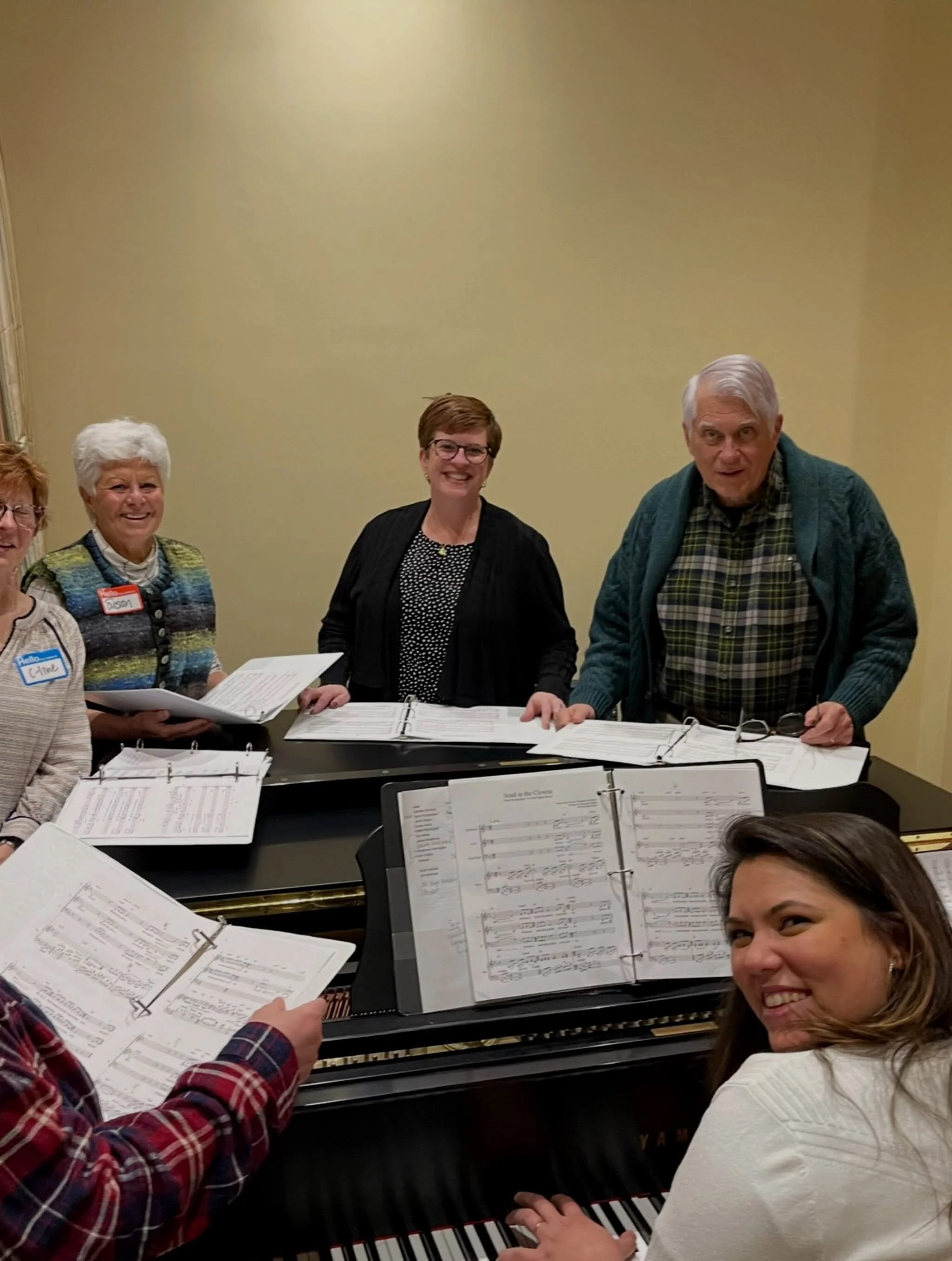 A group of people gathered around a grand piano, with sheet music open on the piano, and some participants holding music sheets, appearing to be participating in a music rehearsal or sing-along event.