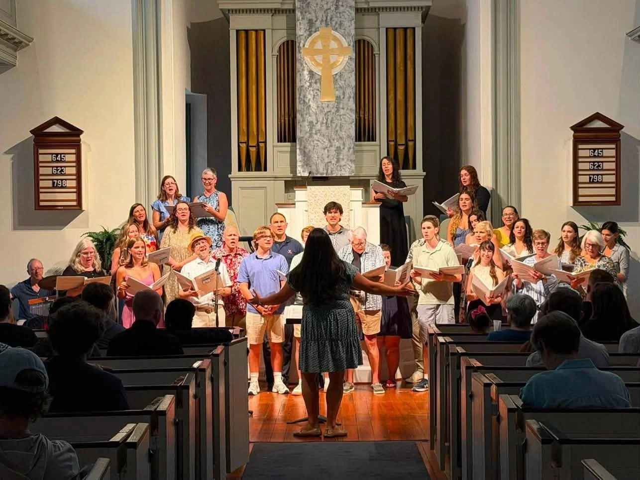 Choir of diverse men and women singing in a church, conducted by a woman standing in front with her back to the camera, with organ pipes and church decor in the background, and congregation seated in the pews.