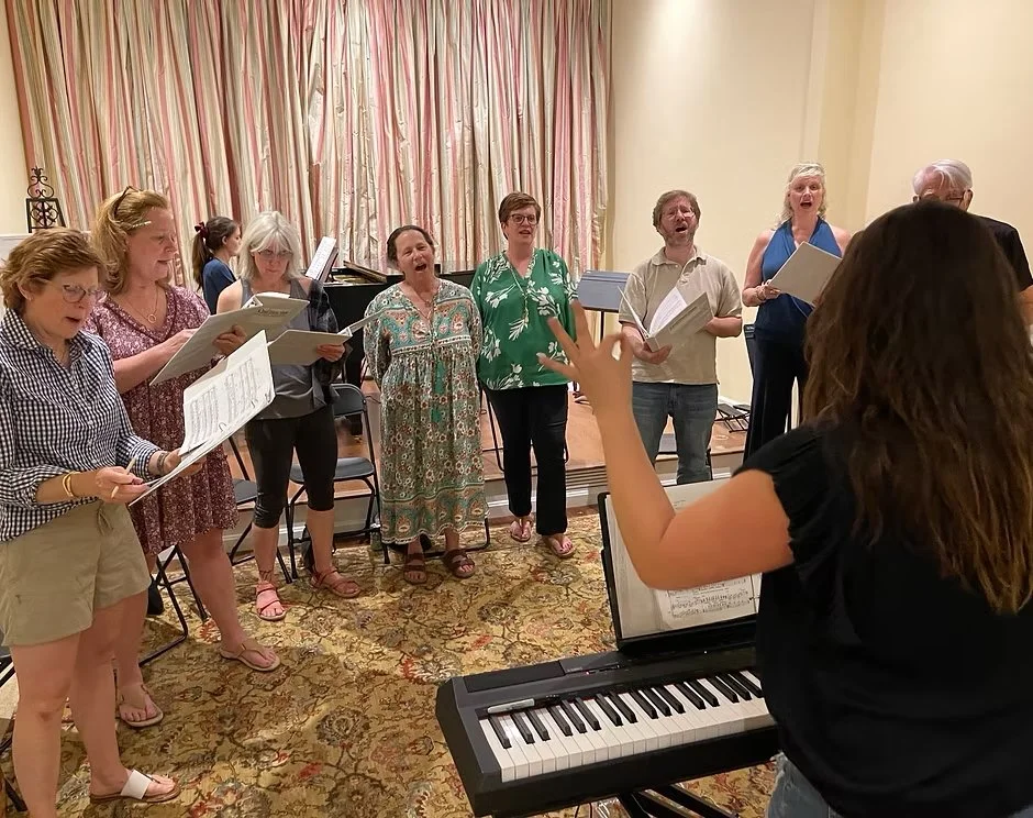 A group of seven adults singing in a choir directed by a woman playing a keyboard in a room with floral carpet and curtains.