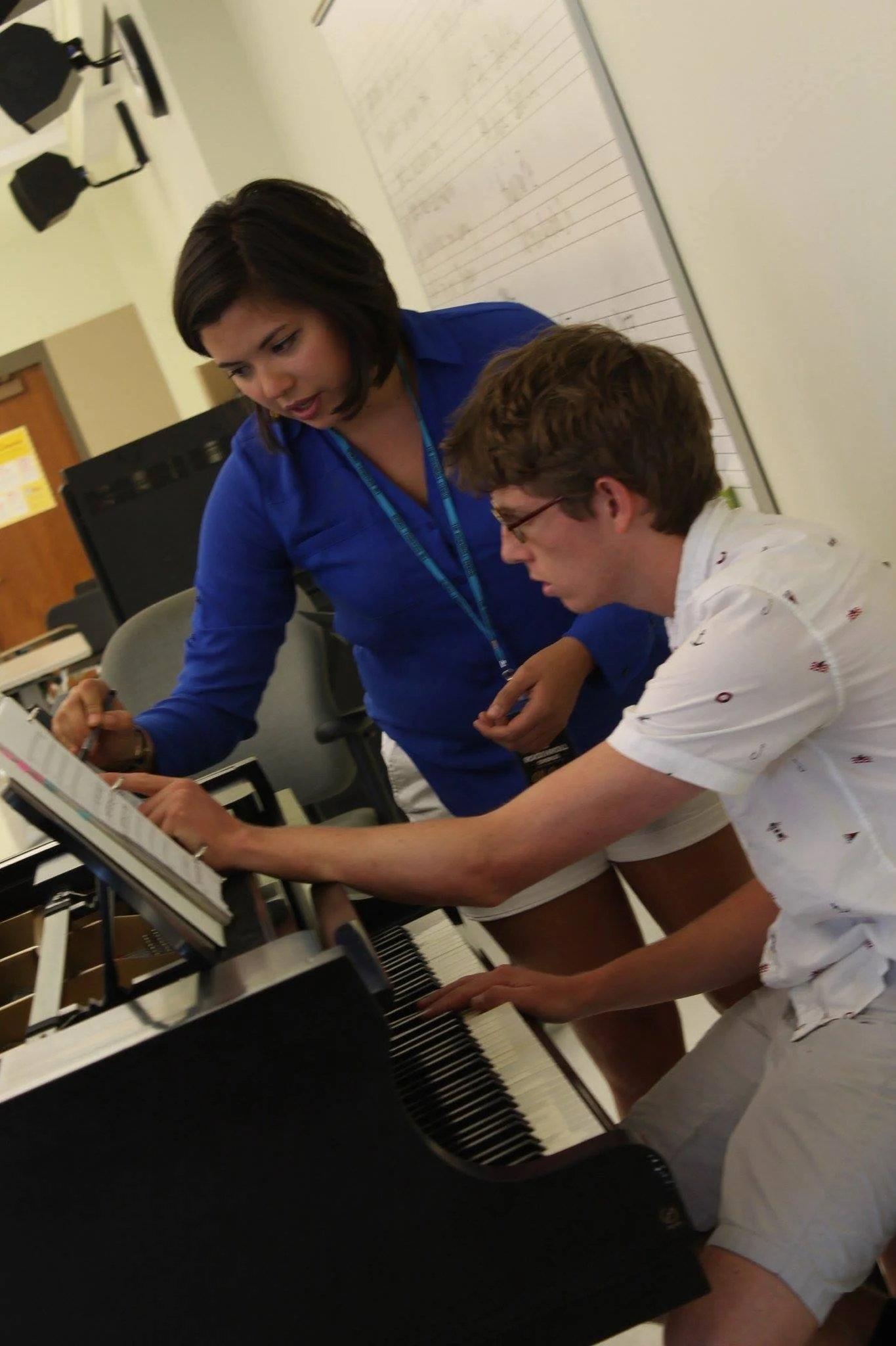 A woman in a blue shirt stands next to a young man in a white shirt with small patterns, who is playing a piano. They are both engaged in the music, with the woman leaning over the young man's shoulder as he focuses on playing the piano.