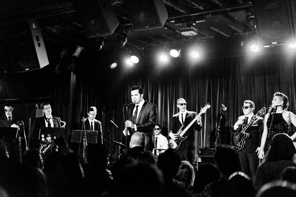 Marianne Cheng singing and playing tambourine with 10-piece, Motown-style band Ernest Ernie and the Sincerities performance at the Bell Hop. 

A black and white photo of a jazz band performing on stage with microphones, instruments, and a curtain bac