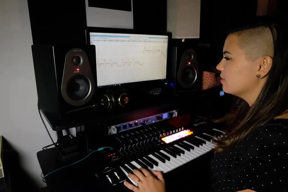 Woman with shaved sides and long brown hair working at a music production station with computer monitors, studio speakers, and a keyboard