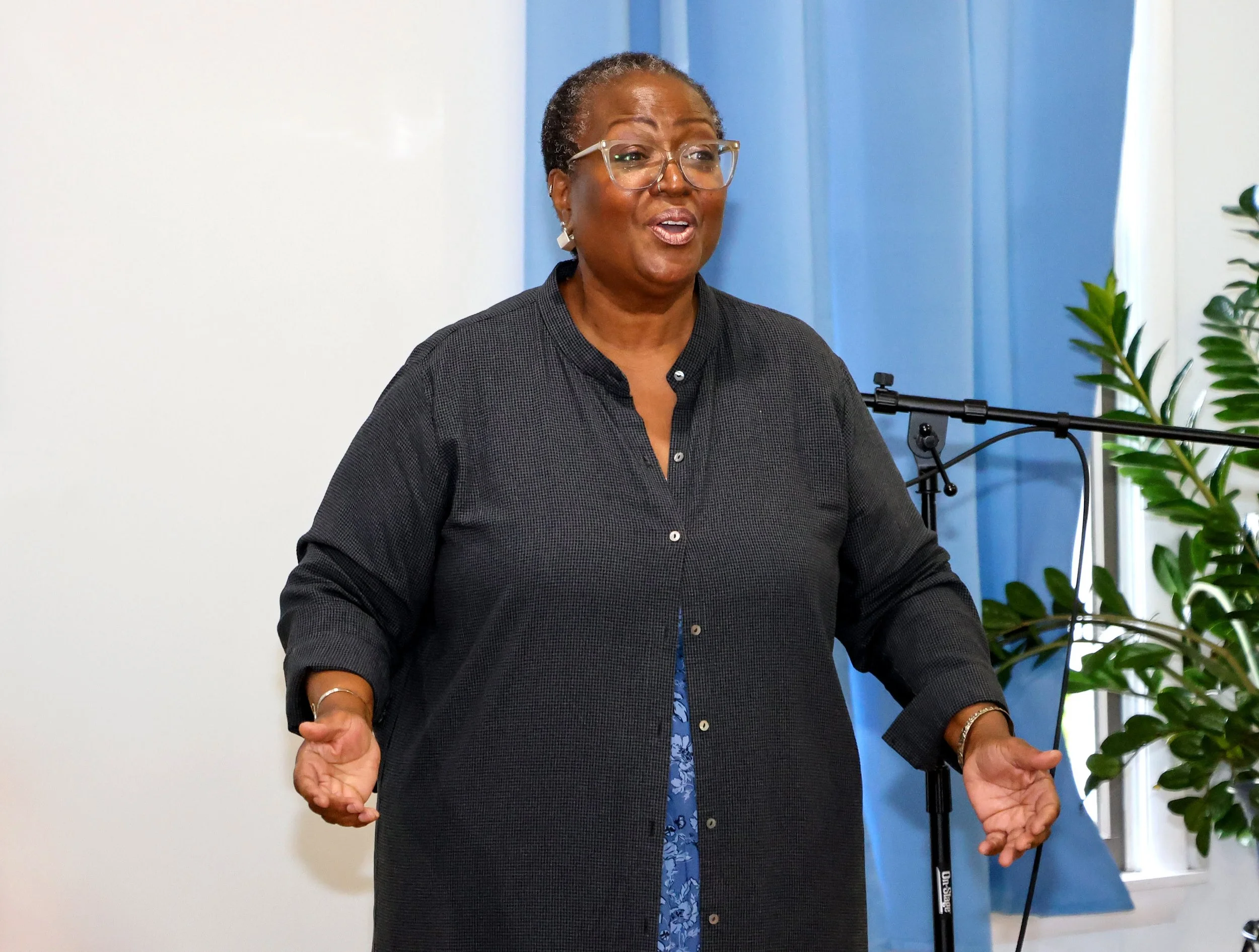 A Black woman speaking or presenting, wearing glasses and a black button-up shirt, with a microphone stand and green plants in the background.