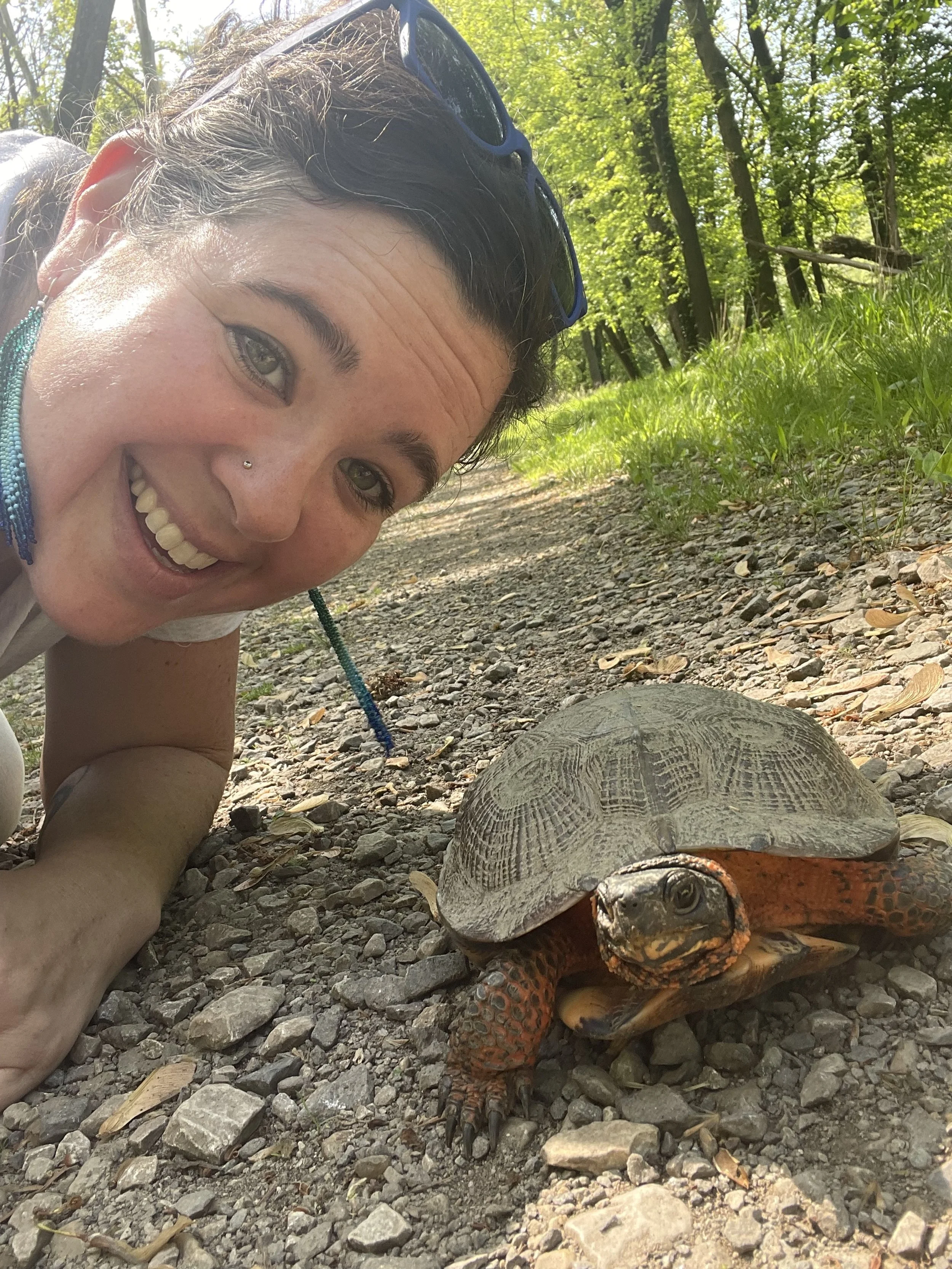 A woman smiling next to a painted turtle on a gravel path in a wooded area.
