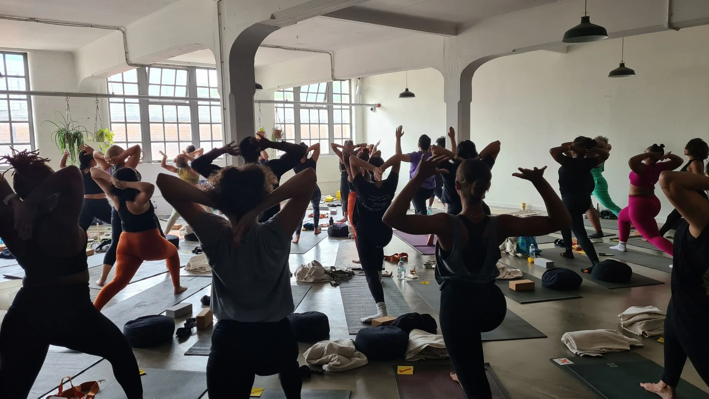 Group of people participating in a yoga class in a bright studio at Space at the Mill with large windows, some people are stretching and doing poses, mats and belongings on the floor.