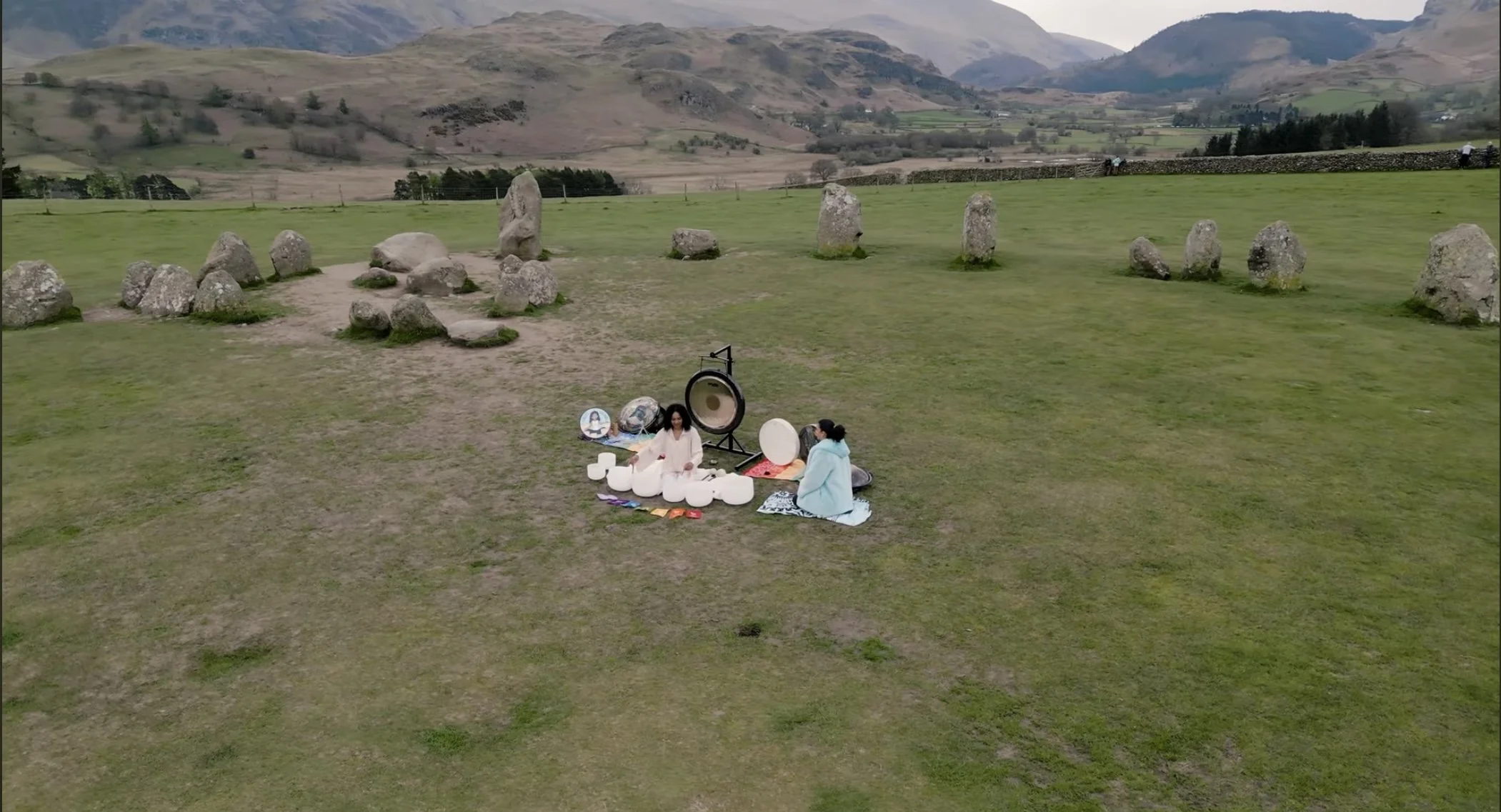 Cinematic drone shot of a women on the grass with crystal singing bowls and other musical instruments, including a large paiste gong, in a grassy field with large stones and a mountainous landscape in the background. Location Castlerigg Stone Cirlce