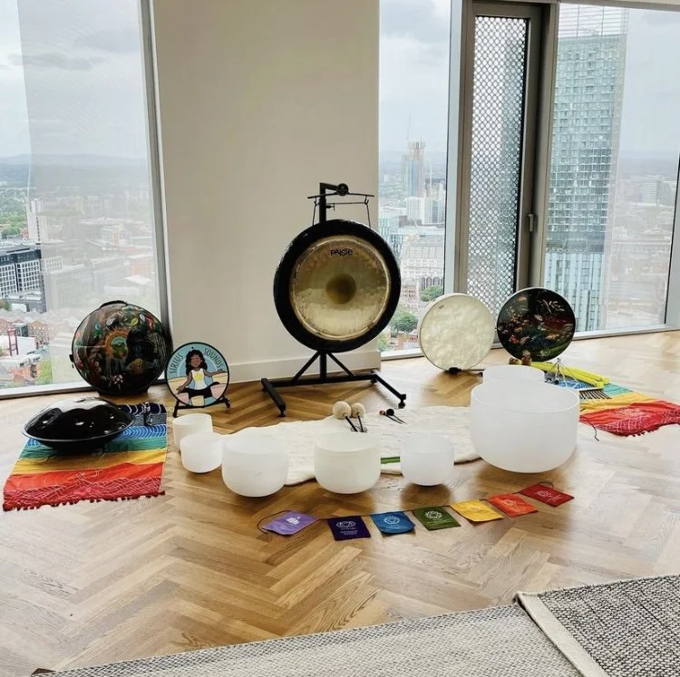 Collection of crystal singing bowls alongside other musical instruments laying on a colorful chakra flag inside high-rise penthouse apartment in with city view in background.