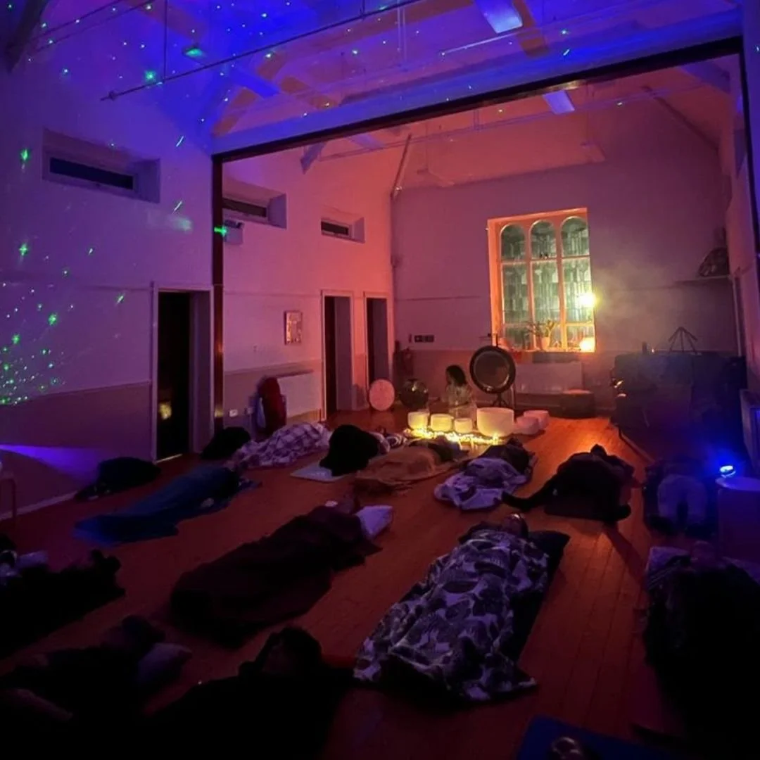 People lying on the floor in a dimly lit room with colorful lights and a large window. Sound bath session in manchester chorlton
