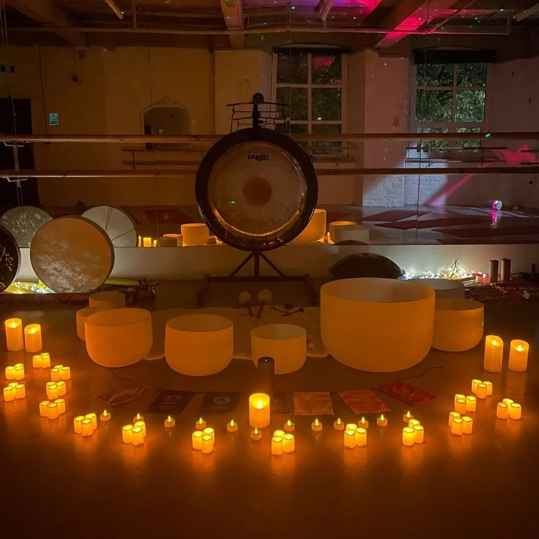 A dimly lit room with a large gong in the center, surrounded by various singing bowls, candles, and decorative objects, creating a calming atmosphere for meditation or sound therapy.