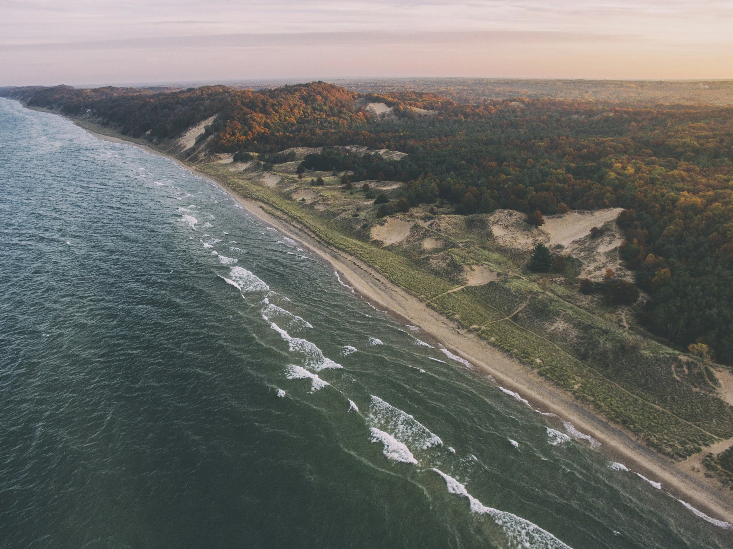 Aerial view of a coastline with waves hitting the sandy beach and a forested area with colorful autumn foliage.