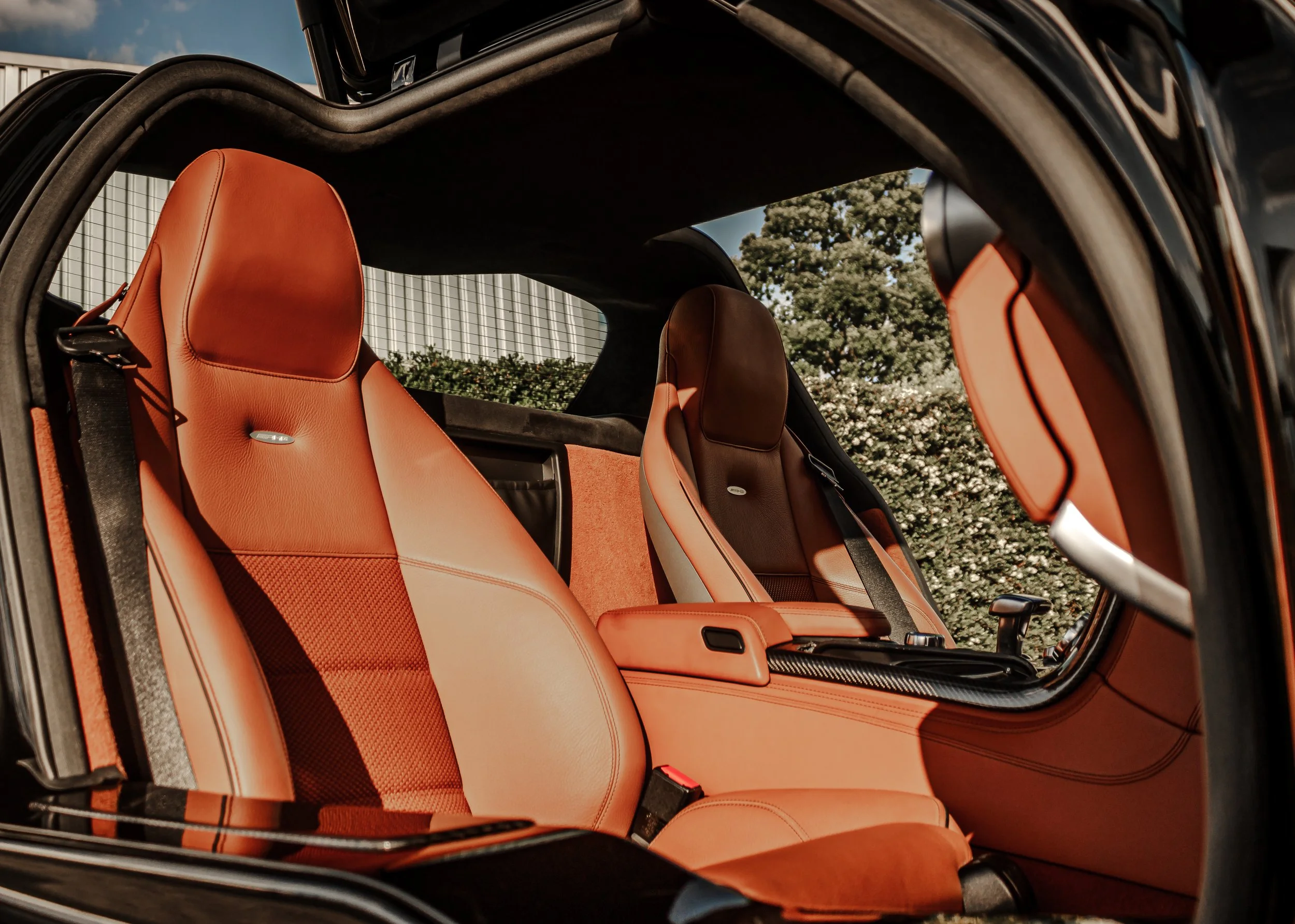 Innensitz eines Autos mit orangefarbenem Leder, Blick durch die offene Tür auf die Sitze und das Autodach, im Hintergrund Bäume und Himmel.