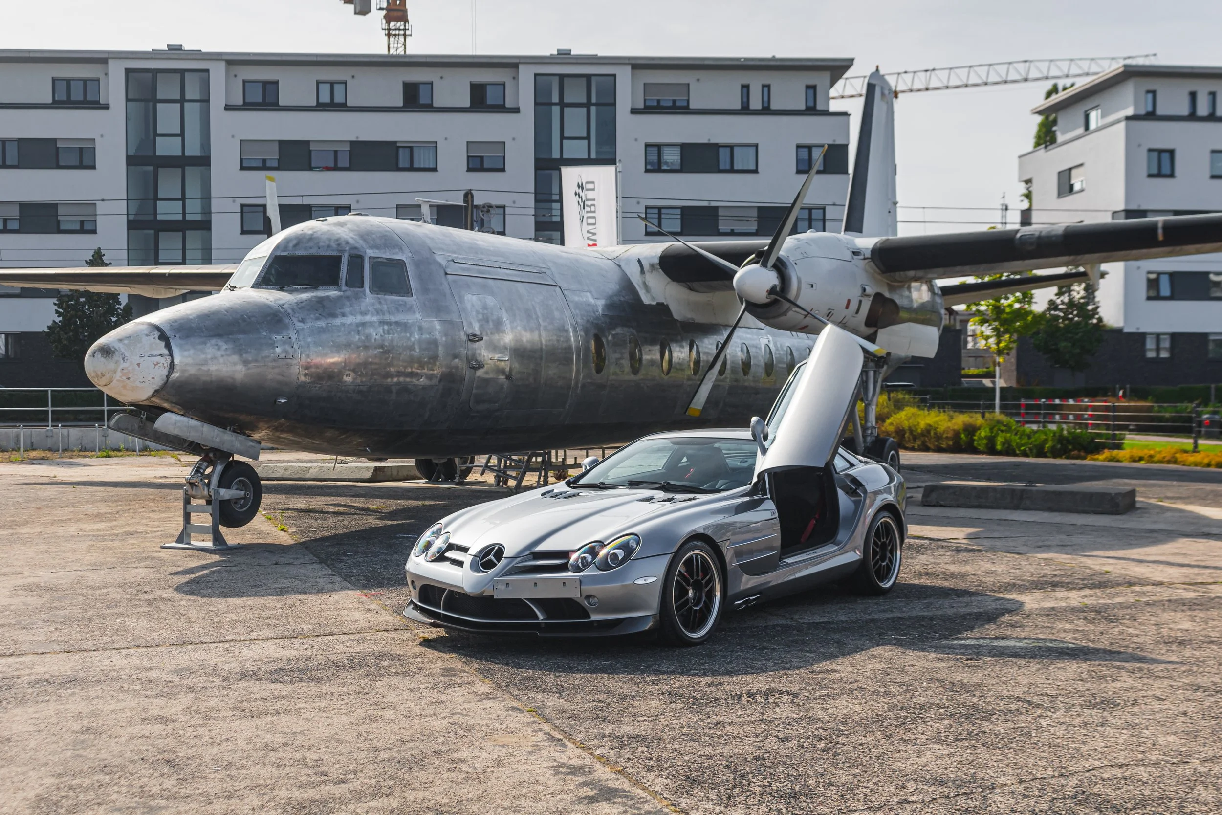 Ein Silber-Sportwagen mit offenen Flügeltüren vor einem alten, abgestellten Flugzeug auf einem Parkplatz in der Stadt.