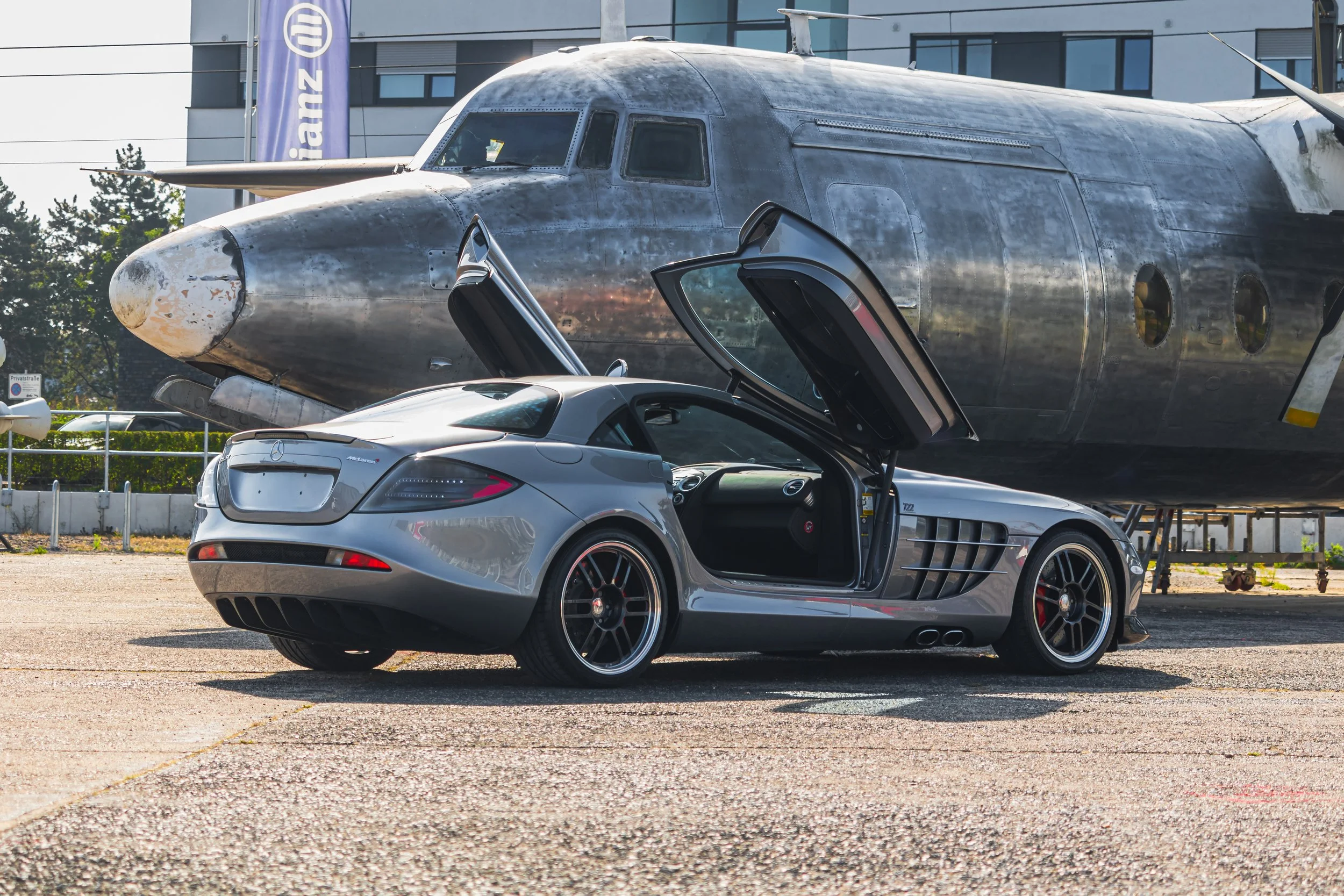 Ein silberner Mercedes-Benz SLS AMG mit Klappflügeln offen vor einem alten Flugzeug aus Edelstahl in einer Flugzeughalle.