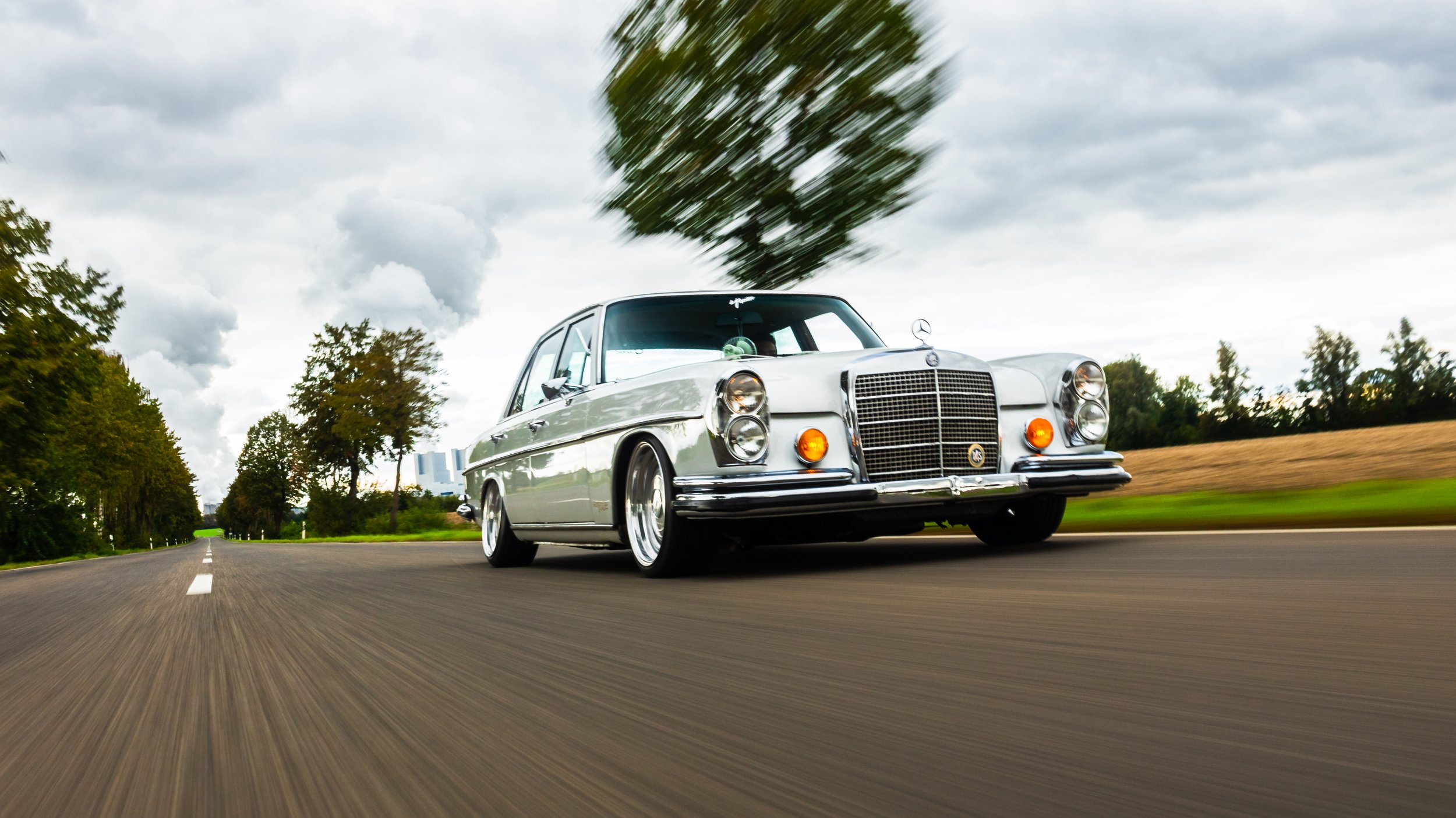 Ein silberner Mercedes-Benz Oldtimer auf einer Landstraße bei bewölktem Himmel, im Vordergrund in Bewegung, mit leichtem Rütteln auf der Straße.