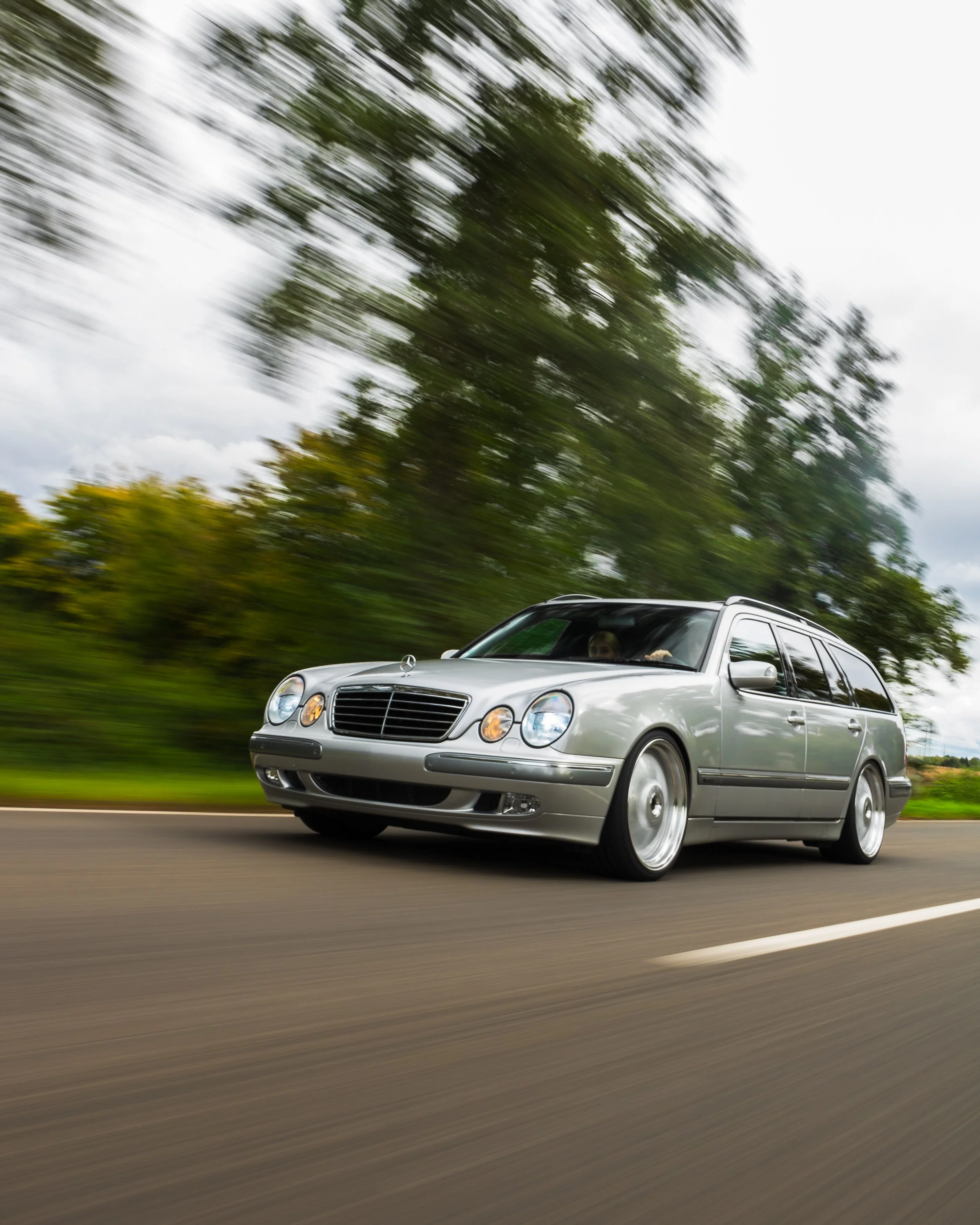 Ein silberner Mercedes-Benz Kombi fährt auf einer Landstraße, während die Umgebung aufgrund der Bewegung unscharf erscheint.