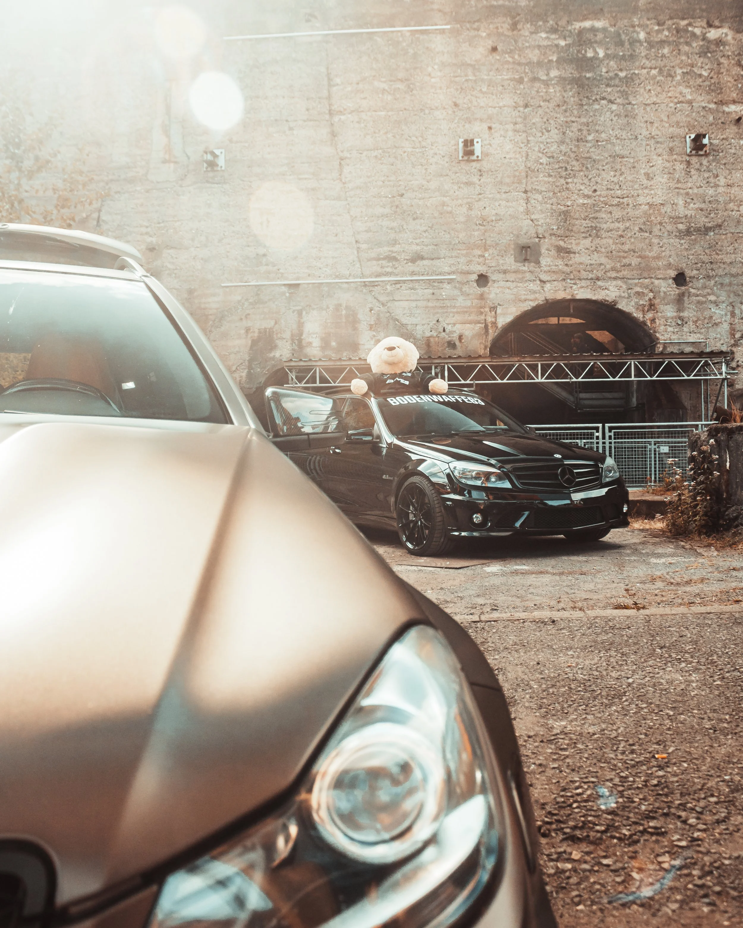 Nahaufnahme der Vorderseite eines C63 im Vordergrund, mit einem schwarzen Mercedes C63  mit Teddy auf dem Dach im Hintergrund, vor einer alten Steinmauer und Baugerüste.
