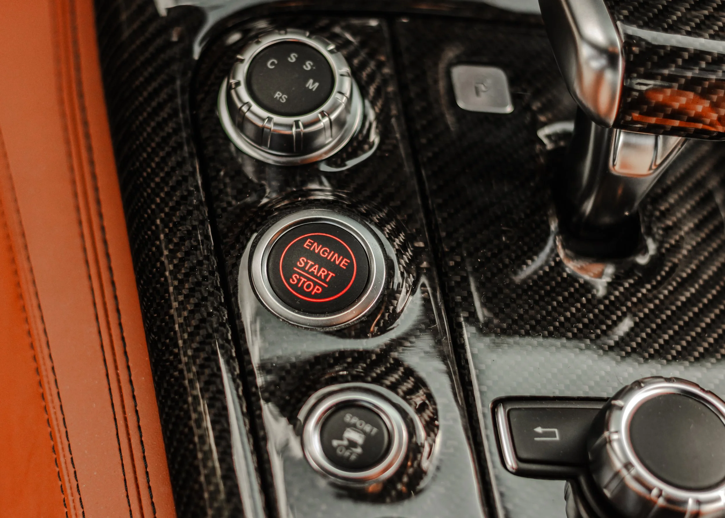 Innenraum eines Autos mit schwarzem Carbon-Design, sichtbar ist ein Start/Stopp-Knopf, ein Drehregler, ein Schalthebel und weitere Bedienelemente.
