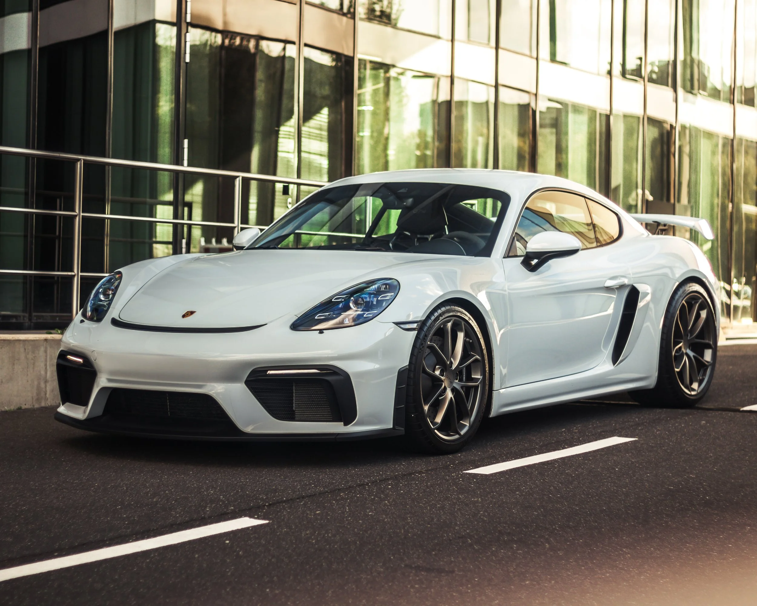 Weißer Porsche 718 Cayman GT4 vor einem modernen Glasgebäude.