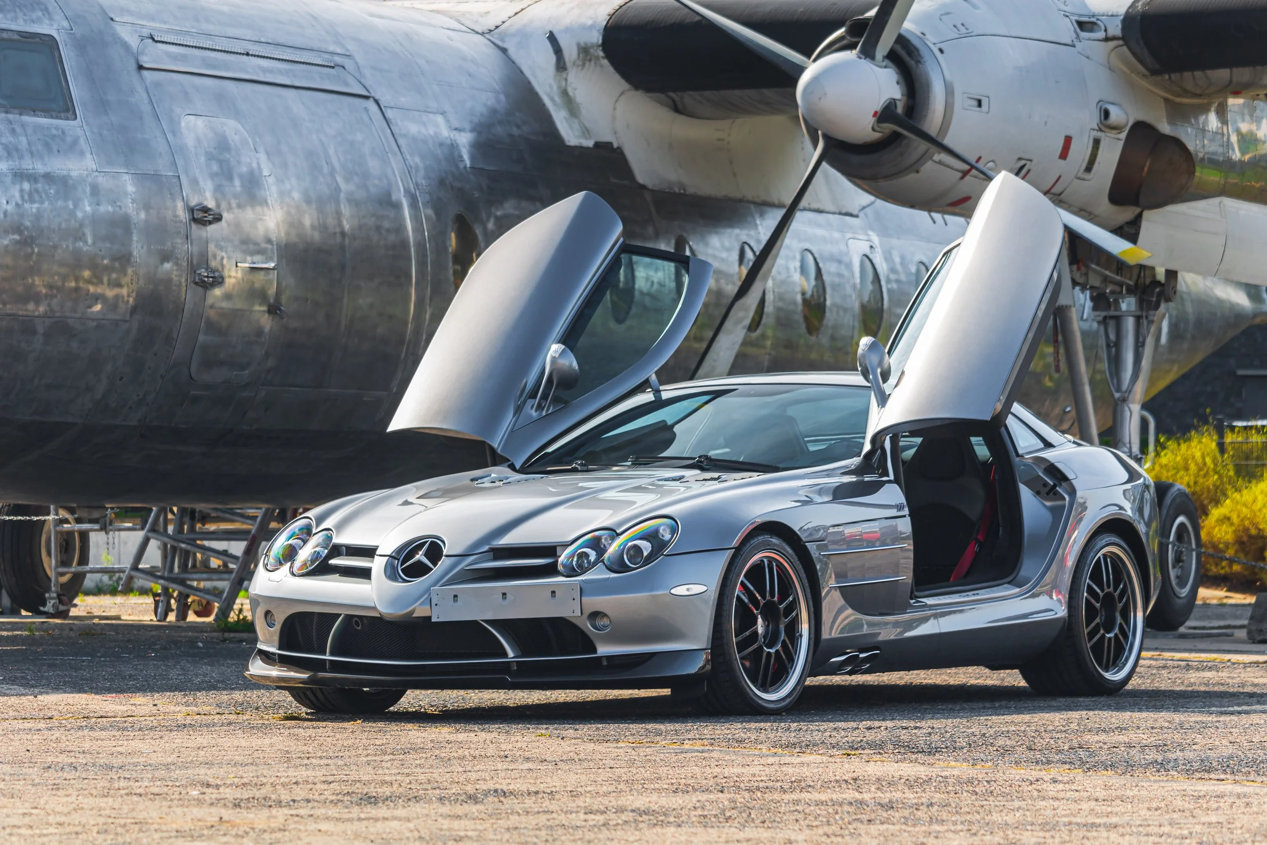 Silberner Mercedes-Benz SLR McLaren mit geöffneten Flügeltüren vor einem alten Flugzeug.