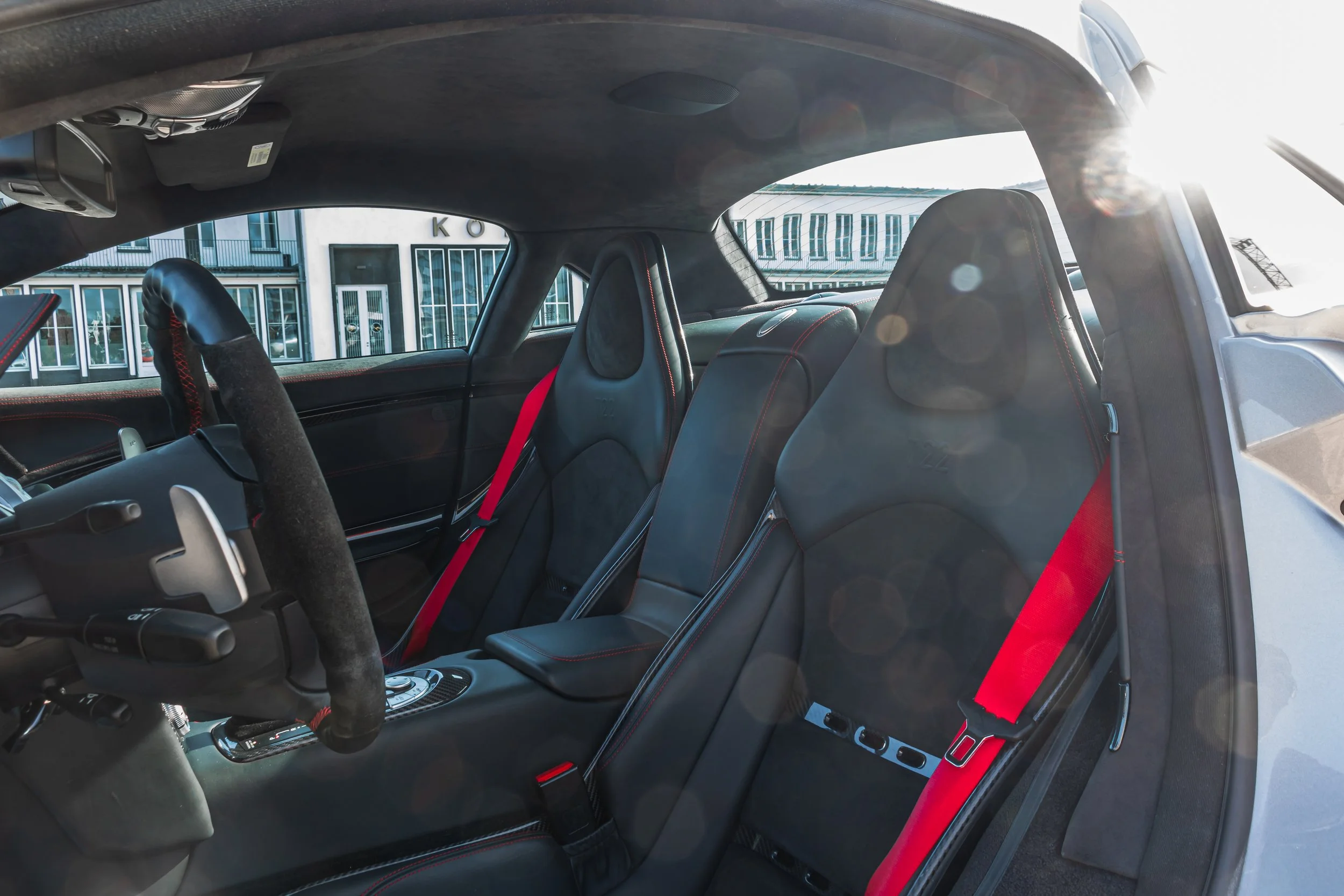 Innenseite eines Autos mit schwarzen Sitzen und roten Sicherheitsgurten, Blick durch die Windschutzscheibe auf ein Gebäude im Hintergrund.
