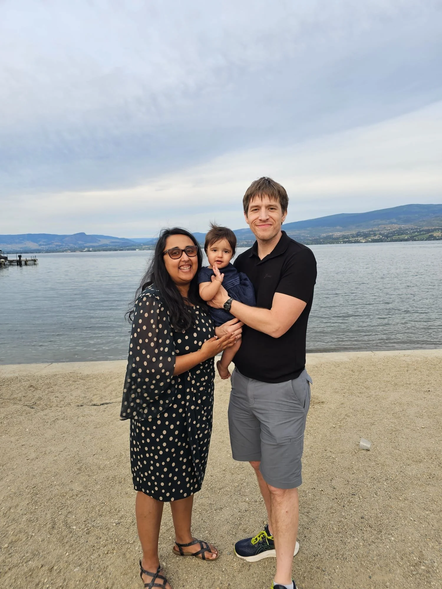 A family of three, a woman, a man, and a toddler, standing on a sandy beach near a body of water with mountains in the background, smiling at the camera.