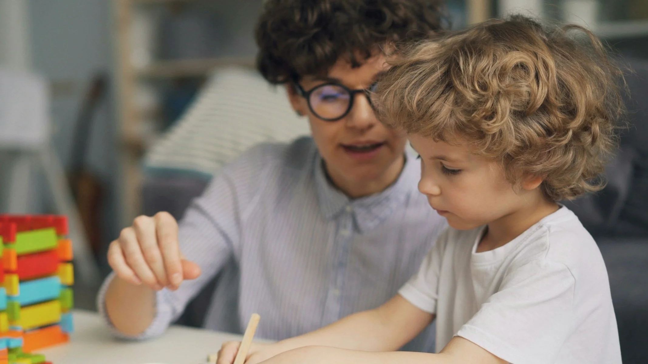 A nanny caregiver male adult with curly hair and glasses helps a young boy with curly hair play a game with colorful wooden pieces made of sensory montessori materials