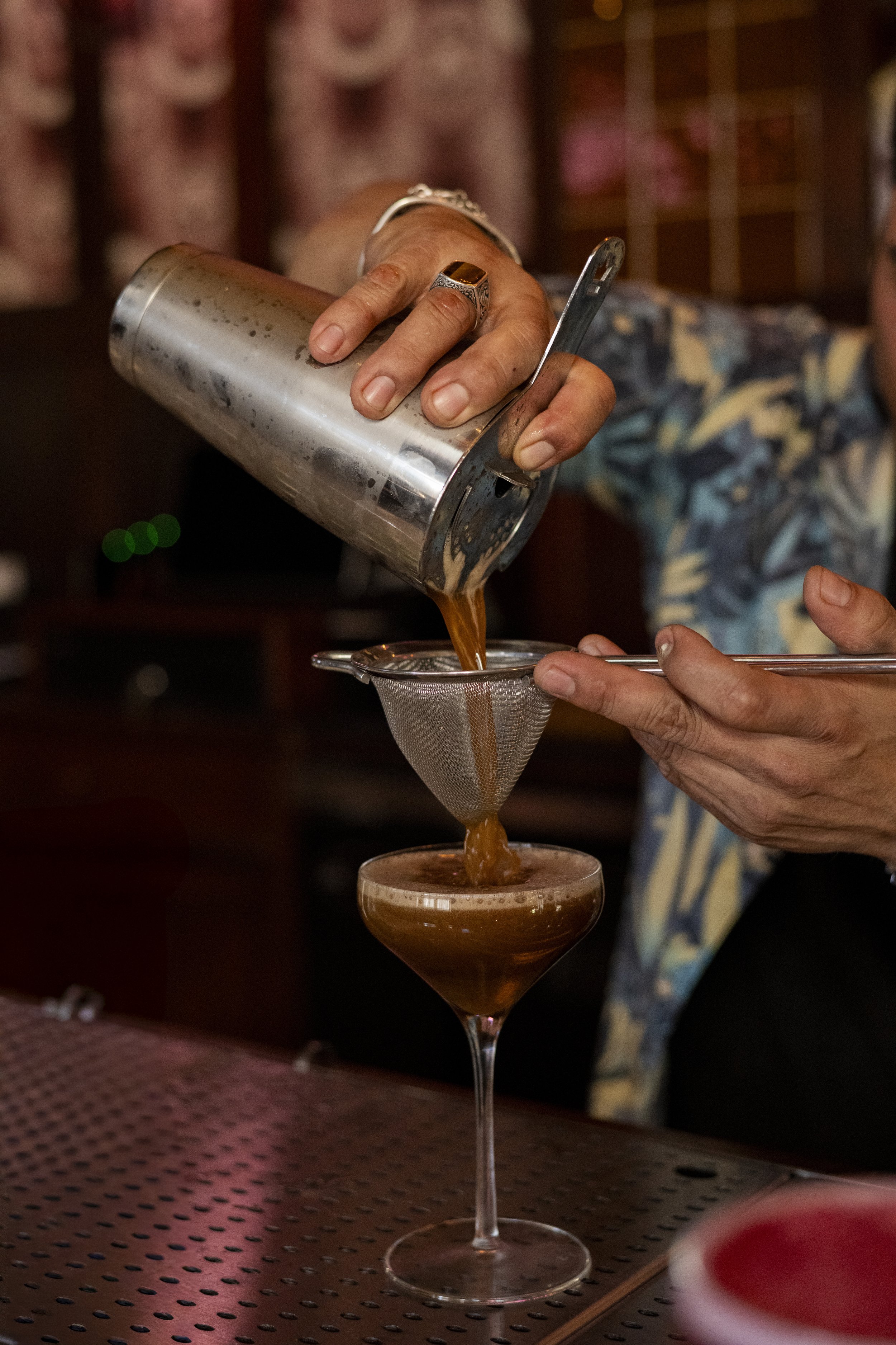 hands pouring brown colored cocktail into tall glass