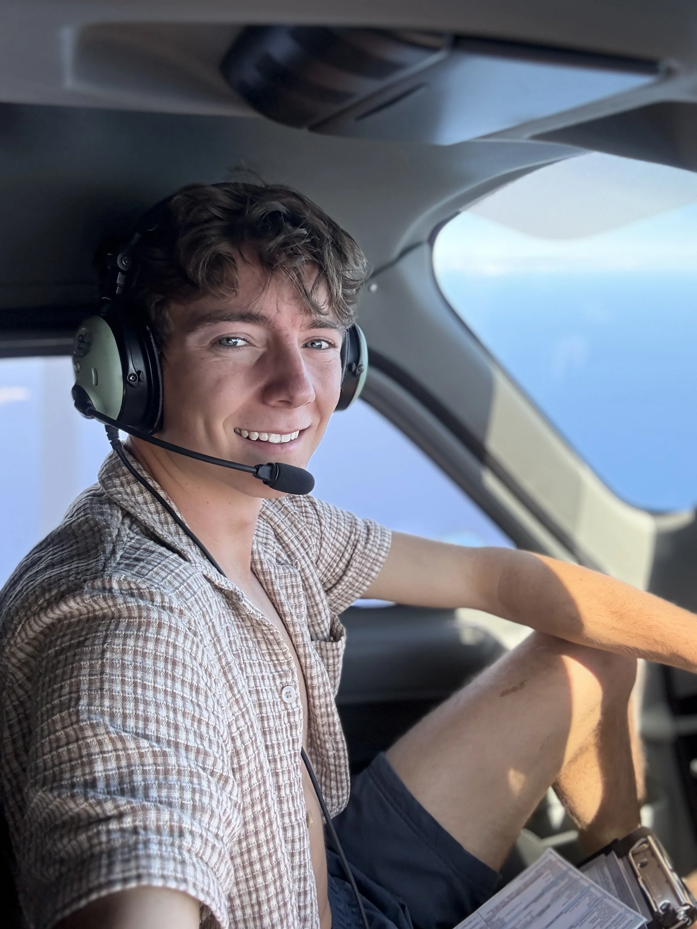 Young male airplane pilot sitting in cockpit, smiling, wearing headset, checklists in hand.