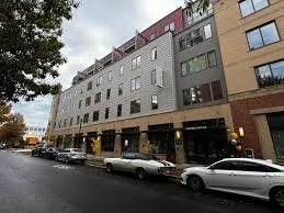 Multistory modern residential building with storefronts on the ground level, parked cars, and cloudy sky.