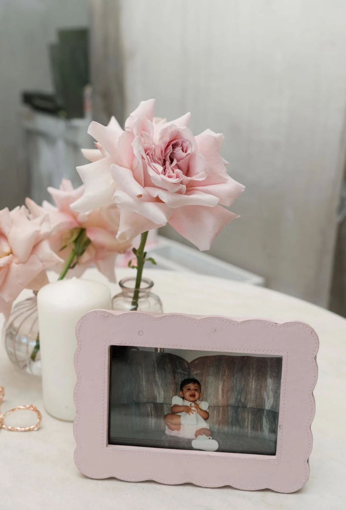A pink framed photo of a happy baby sitting on a couch, with a pink flower bouquet in a glass vase and a white candle on a table behind it.