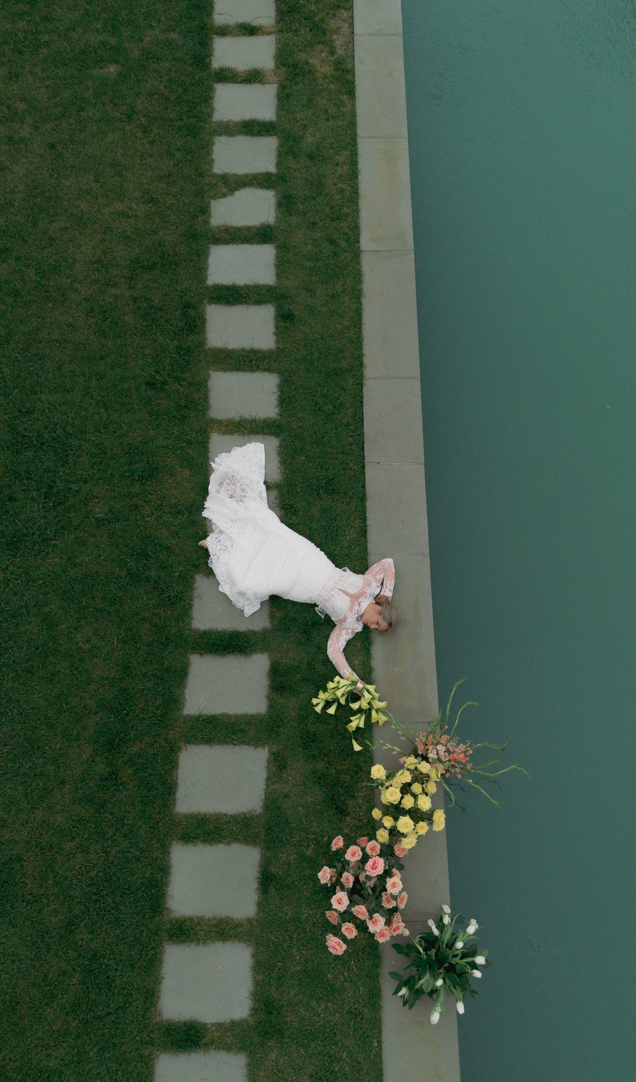 A woman in a white wedding dress lying on the grass next to a large bouquet of flowers, with a pool or water feature nearby.