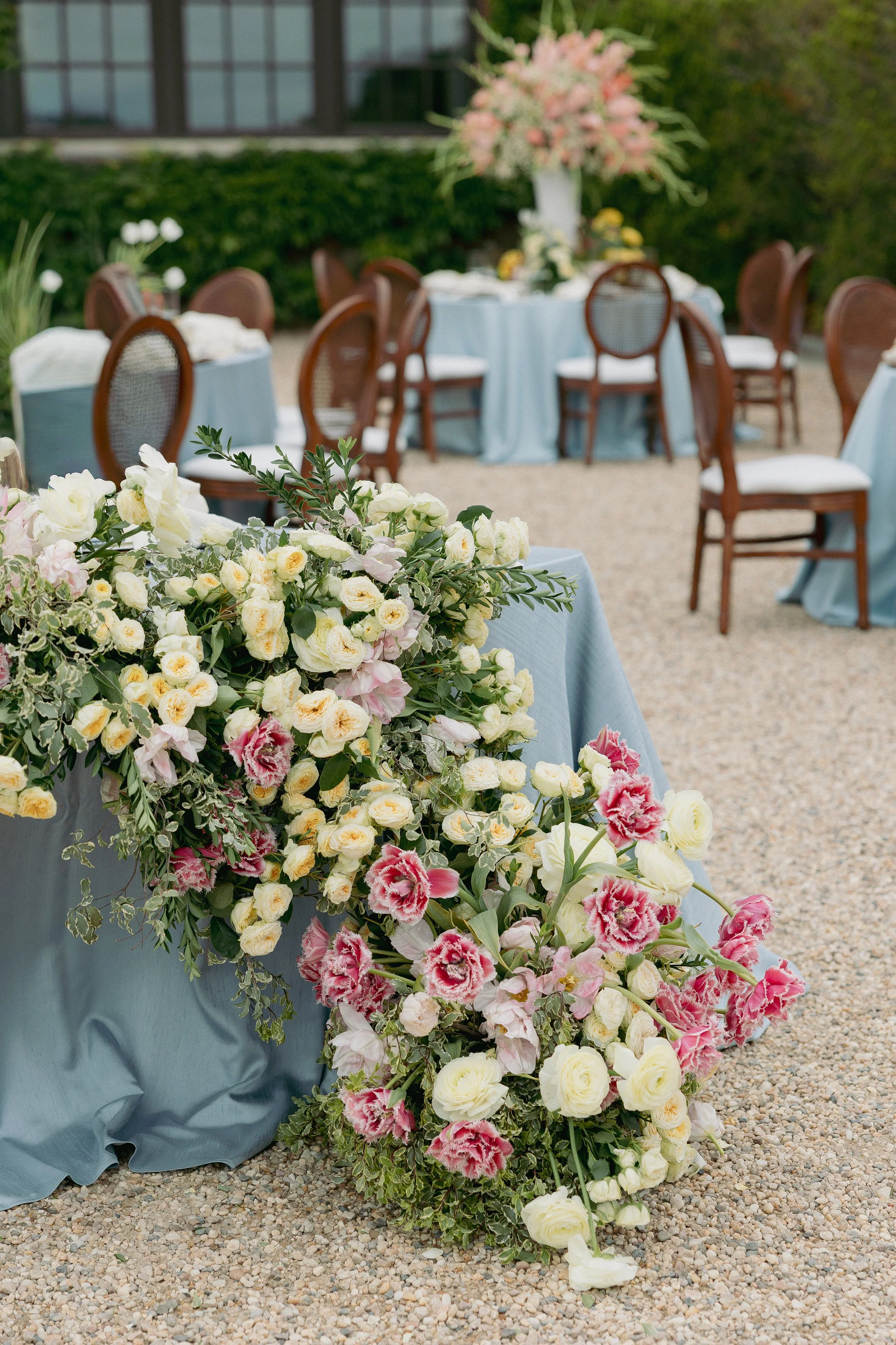A large floral arrangement with white, pink, and cream roses, along with green foliage, is placed on a table covered with light blue fabric at an outdoor event. In the background, there are multiple tables with similar light blue tablecloths and wooden chairs, set in a garden or yard with green bushes and trees.