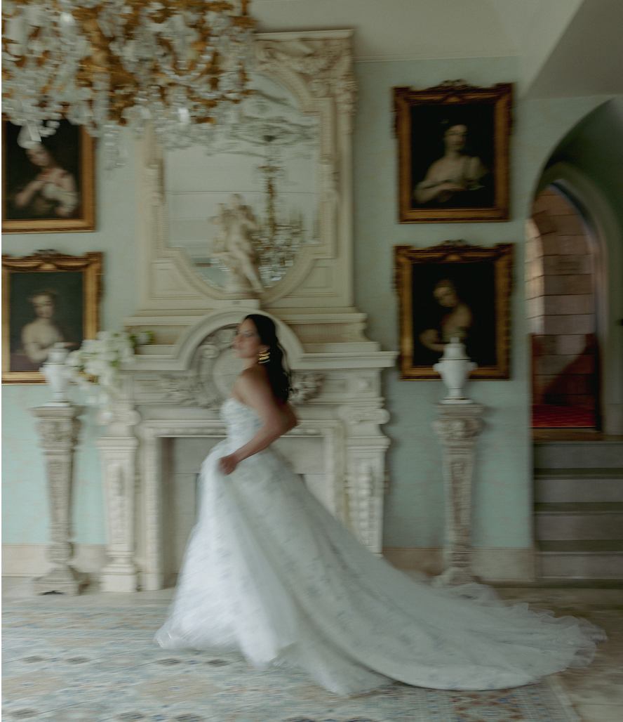 A woman in a wedding dress standing in an elegant, vintage-style room with ornate furniture and framed portraits on the wall.